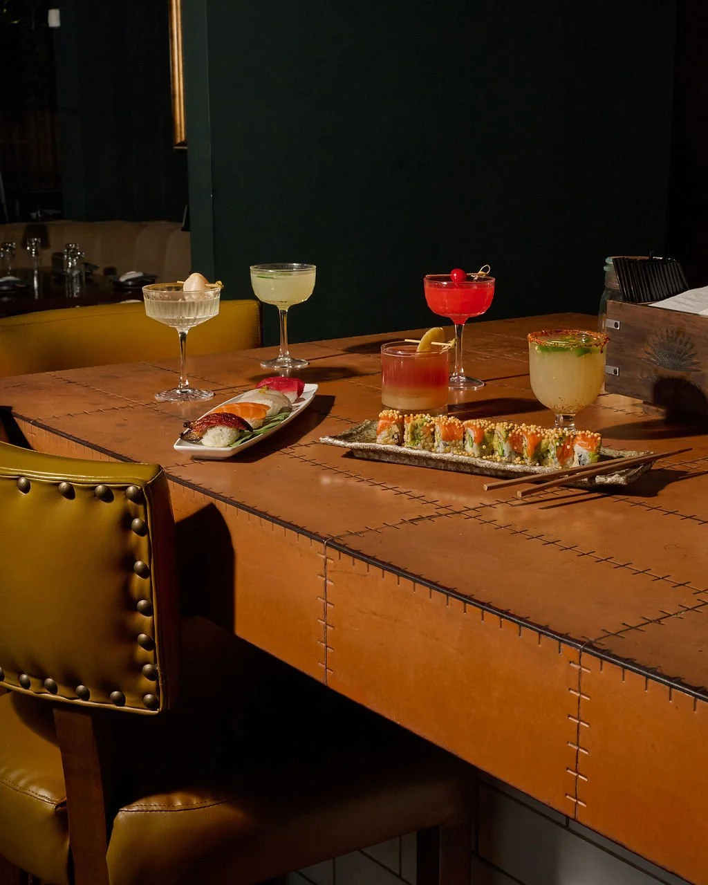 A wooden table set with five colorful cocktails and a plate of sushi rolls in a dimly lit restaurant.