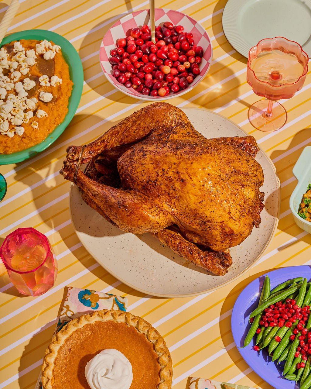 A roasted turkey on a white plate with side dishes including cranberry sauce, green beans with cranberries, sweet potato casserole with popcorn marshmallows, pumpkin pie with whipped cream, and a bowl of fresh cranberries, all arranged on a yellow and white striped tablecloth.