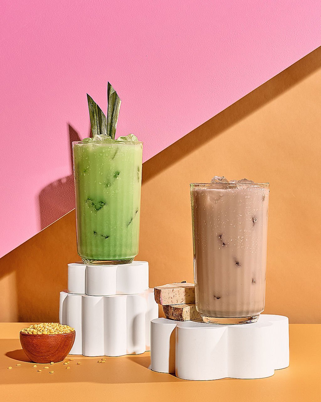 Two glasses of iced beverages, one green and one purple, on white decorative stands against a pink and orange background with small yellow snack in a wooden bowl.