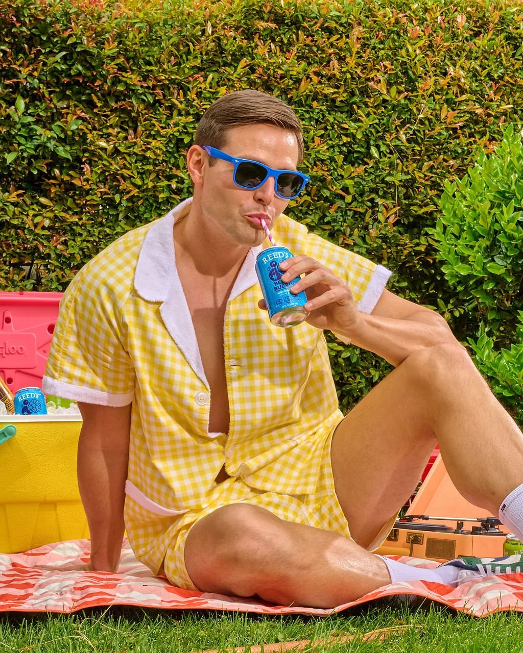 A man with short brown hair wearing blue sunglasses and a yellow checkered short-sleeve shirt is sitting on a grass lawn, sipping a blue soda from a can with a straw. There is a pink cooler, some drinks, and a record player nearby, with a hedge in the background.