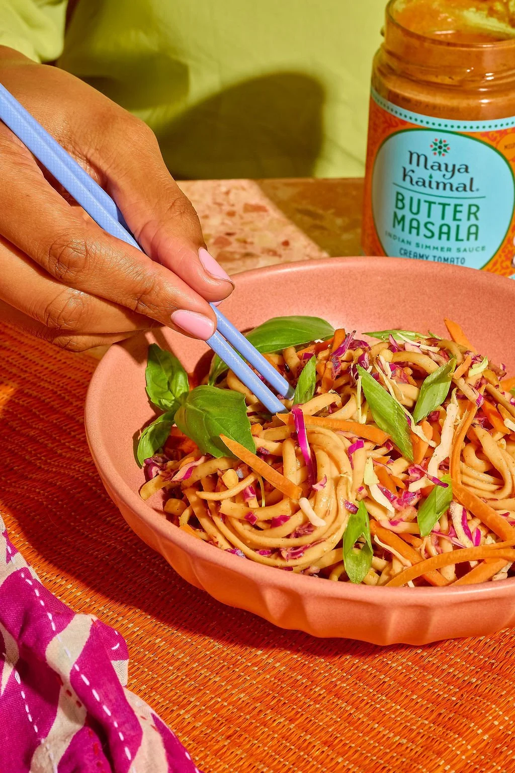 A person using blue chopsticks to eat a colorful noodle salad garnished with basil leaves, with a jar of Maya Kaimal butter masala sauce in the background.