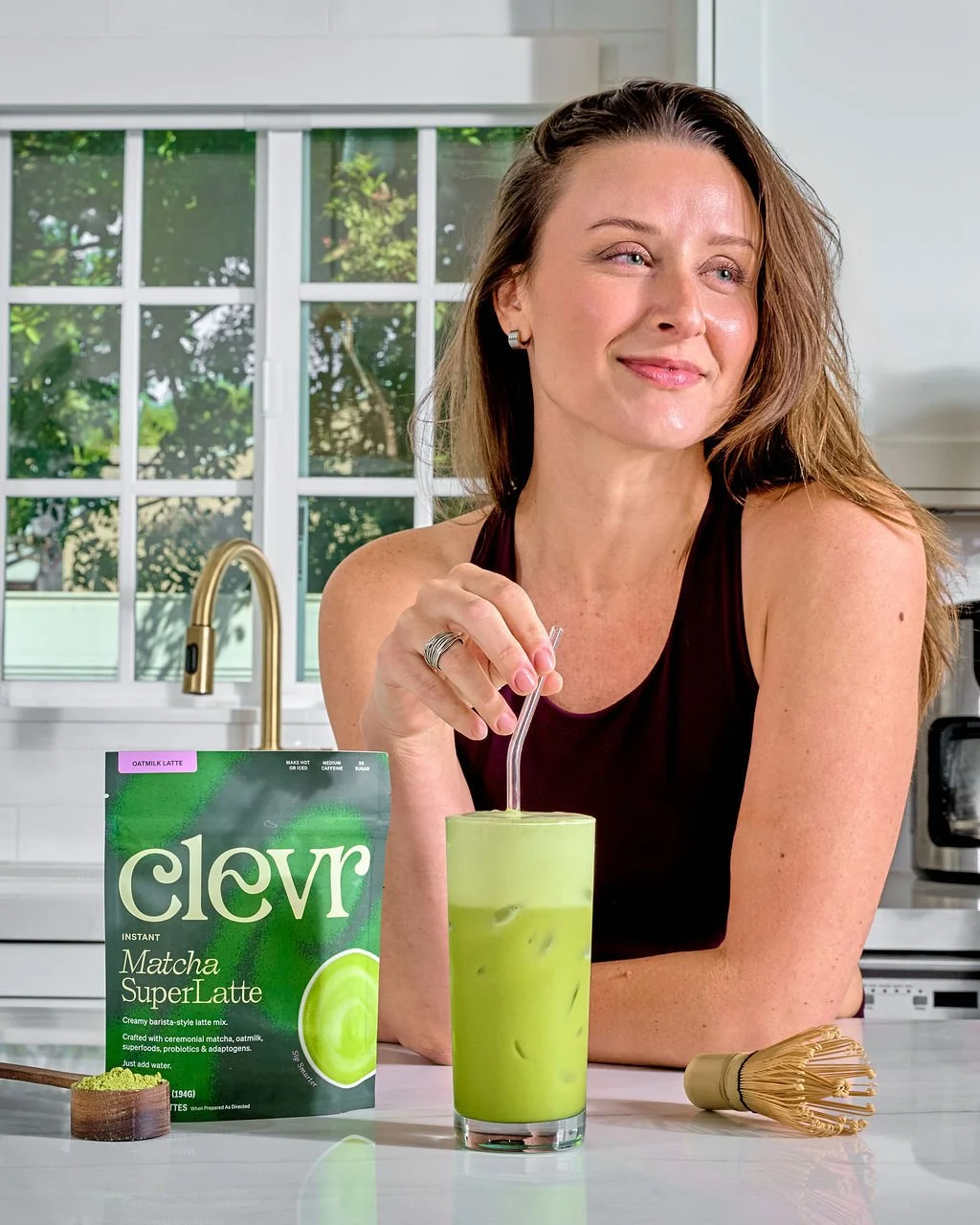 A woman with brown hair wearing a black sleeveless top, sitting at a white kitchen counter with a green matcha latte and a packet of Clever Matcha SuperLatte in front of her. She is smiling, holding a straw in her drink, with a bamboo whisk on the co