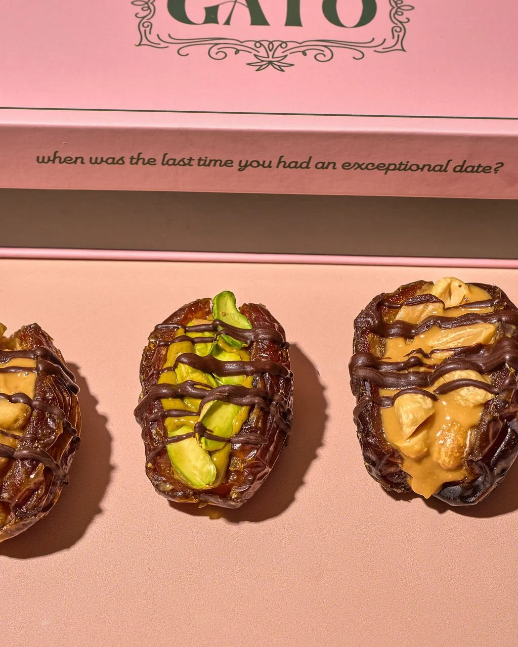 Three chocolate-dipped dates filled with nuts and topped with various toppings, placed on a pink surface with a pink box that has the question 'when was the last time you had an exceptional date?' written on it.