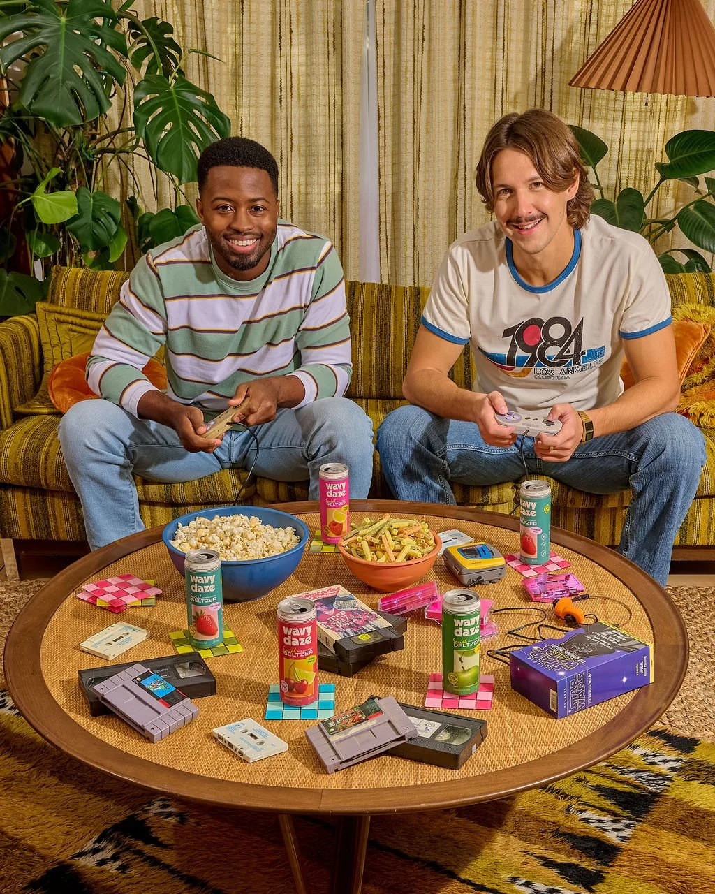 Two men sitting on a yellow striped sofa playing video games, surrounded by snacks, soda cans, and retro gaming cartridges on a wooden table.