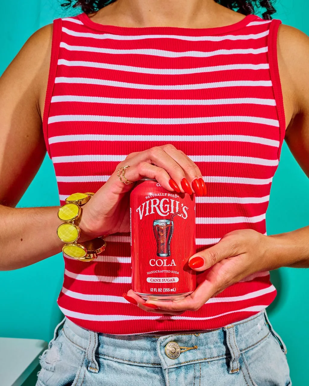 Person in a red and white striped sleeveless top holds a red can of Virgils Cola, standing against a blue background. The person has painted red nails, wears a yellow bracelet, a gold ring, and light blue jeans.