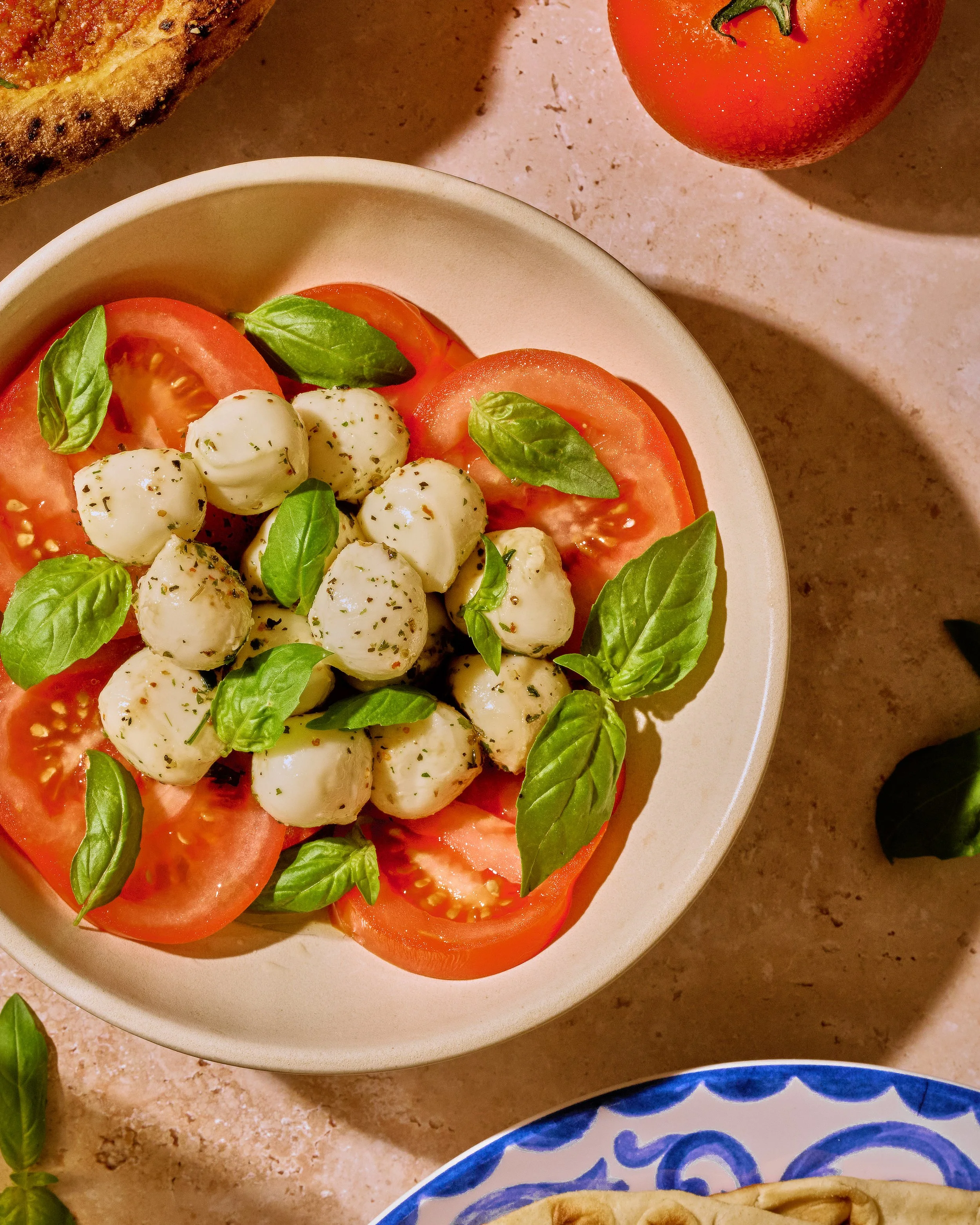 Caprese salad with sliced tomatoes, fresh mozzarella balls, basil leaves, and black pepper in a white bowl on a beige countertop. Part of a toasted bread slice and a tomato are visible in the background.