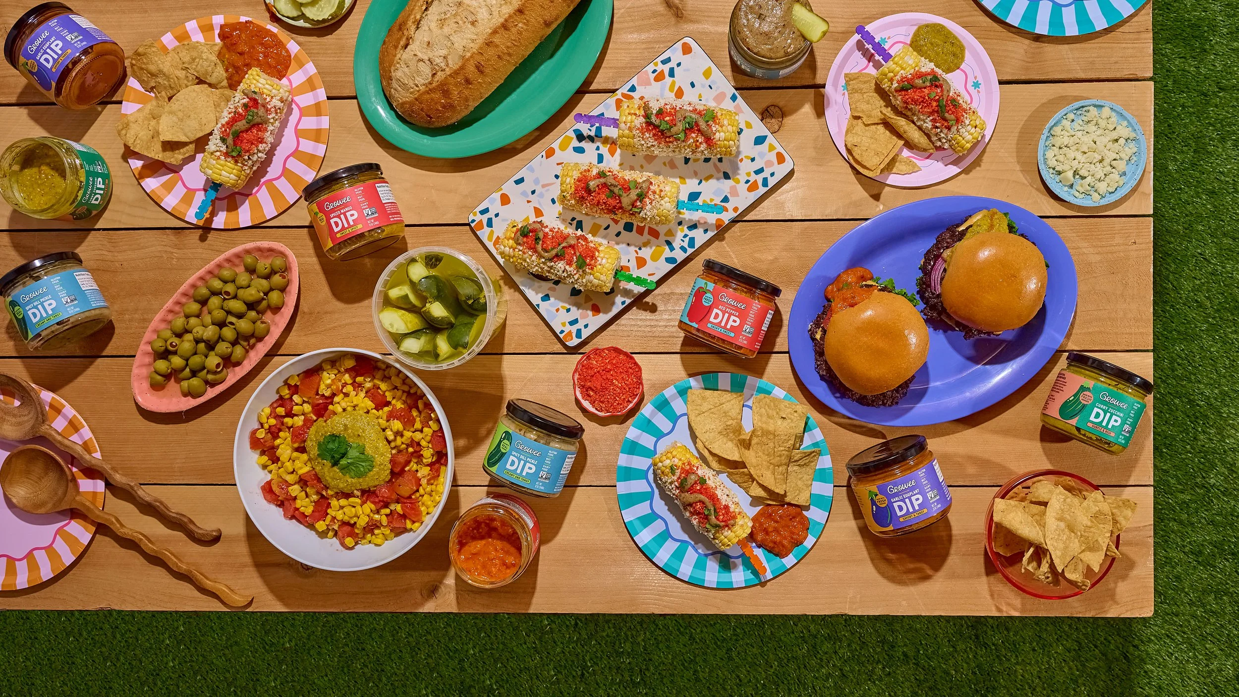 A variety of colorful Mexican dishes and snacks on a wooden table, including corn on the cob with chili and seasoning, burgers with buns, bowls of guacamole, chopped vegetables, and multiple jars of salsa and dips.