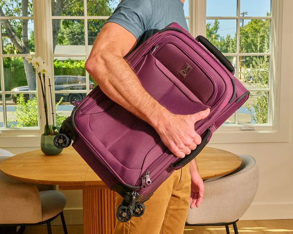 Person holding a purple rolling suitcase inside a bright room with large windows, a wooden table, and chairs.