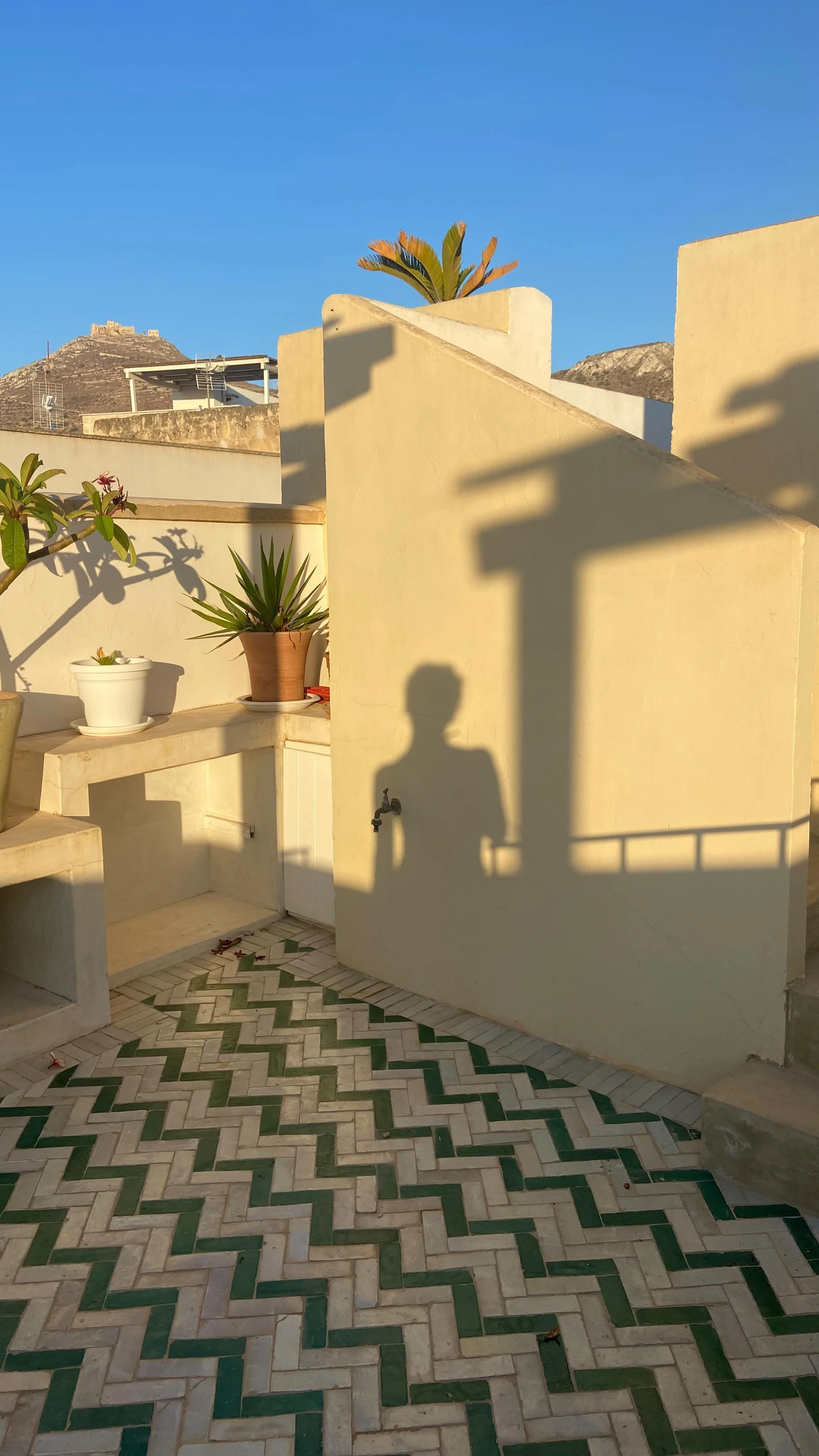 Sunlit outdoor terrace with potted plants on built-in counters, patterned tile floor, and shadows of a lamppost and person cast on a beige wall, with distant hills and a historical fortress under a clear blue sky.