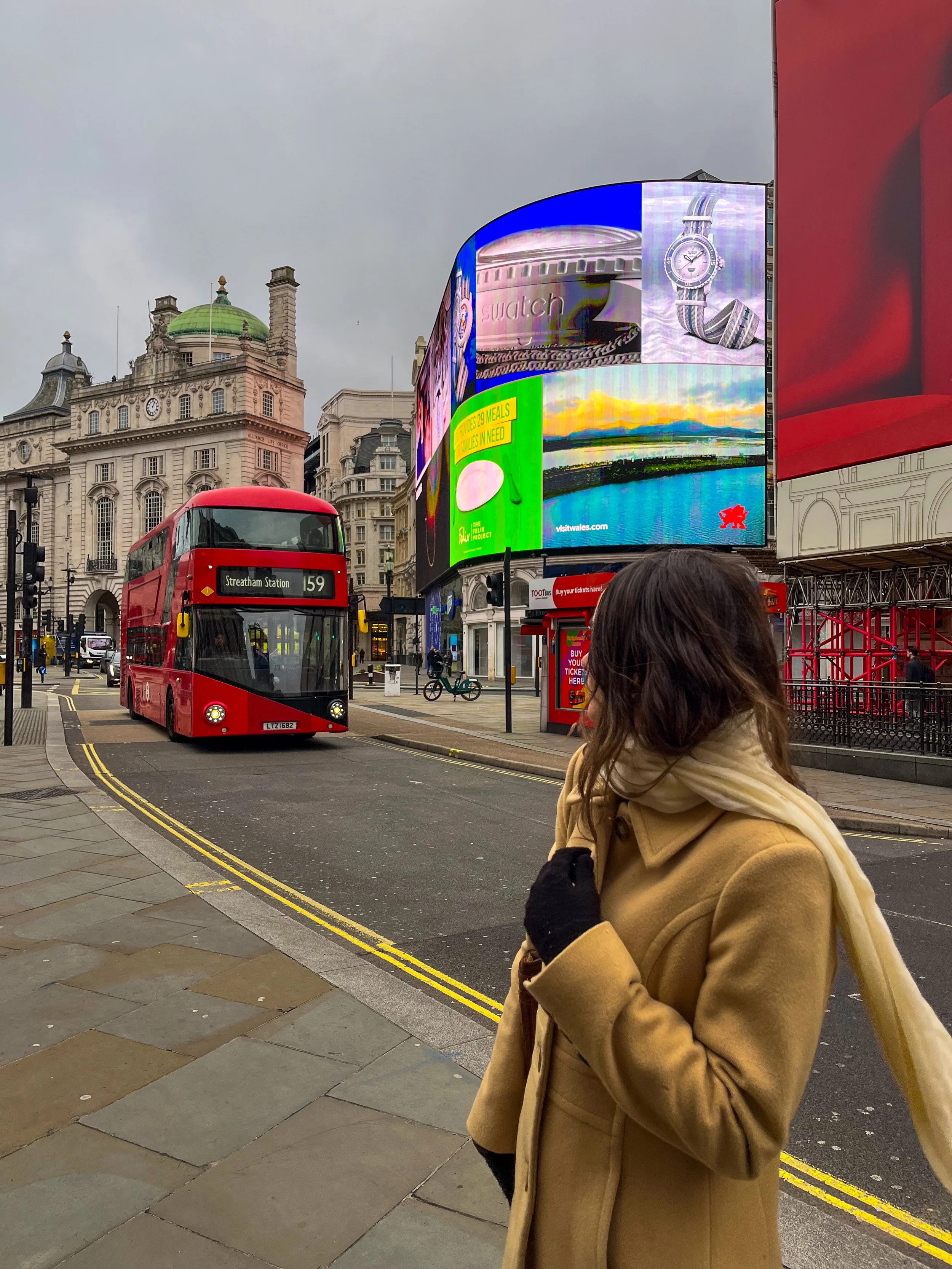 A woman with brown hair wearing a tan coat and a cream scarf standing on a city sidewalk in front of digital billboards, a red double-decker bus on the street, and historic buildings in the background, under a cloudy sky.