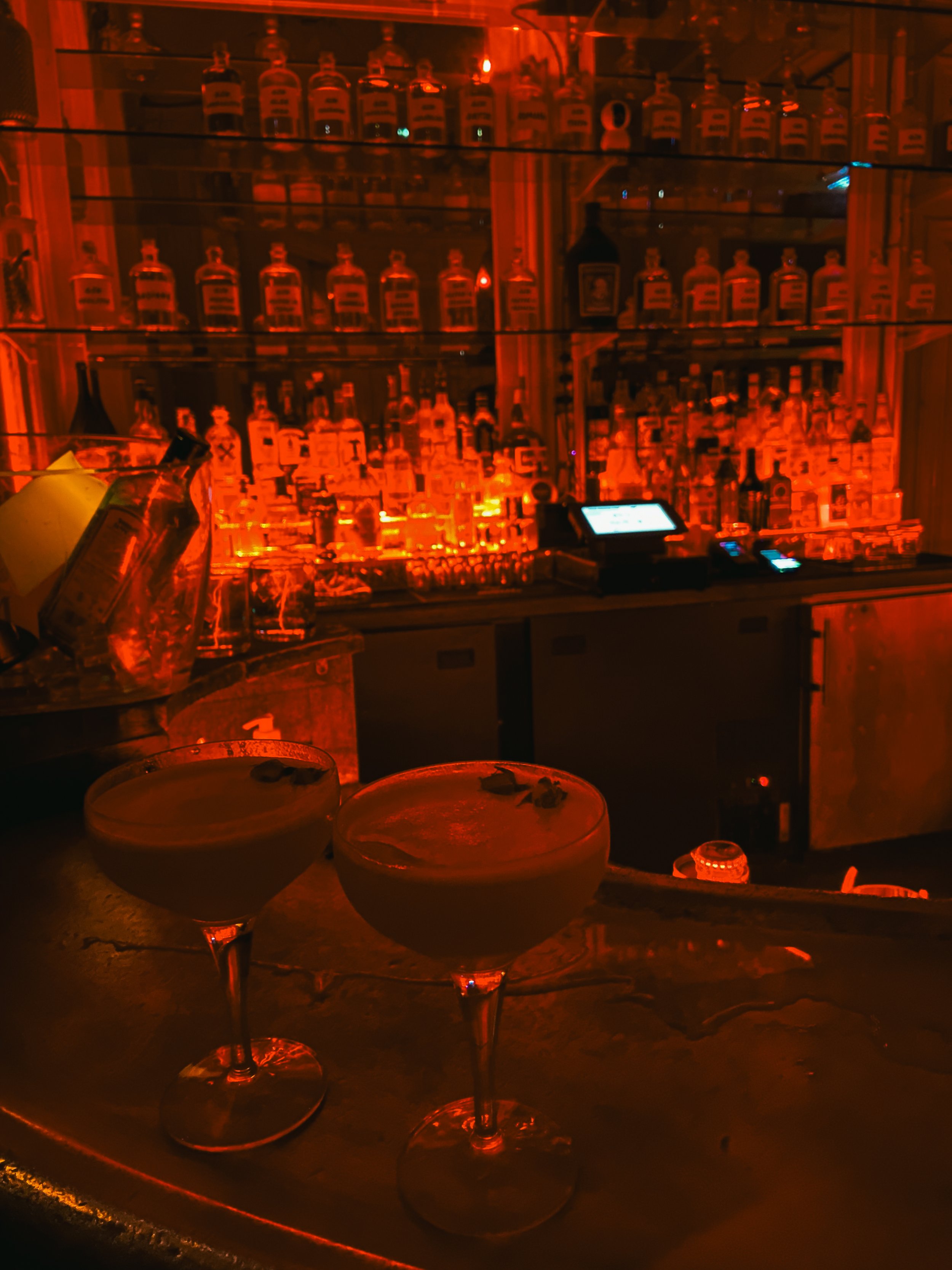 Two cocktail glasses on a bar counter in a dimly lit bar with a brightly lit back bar stocked with bottles