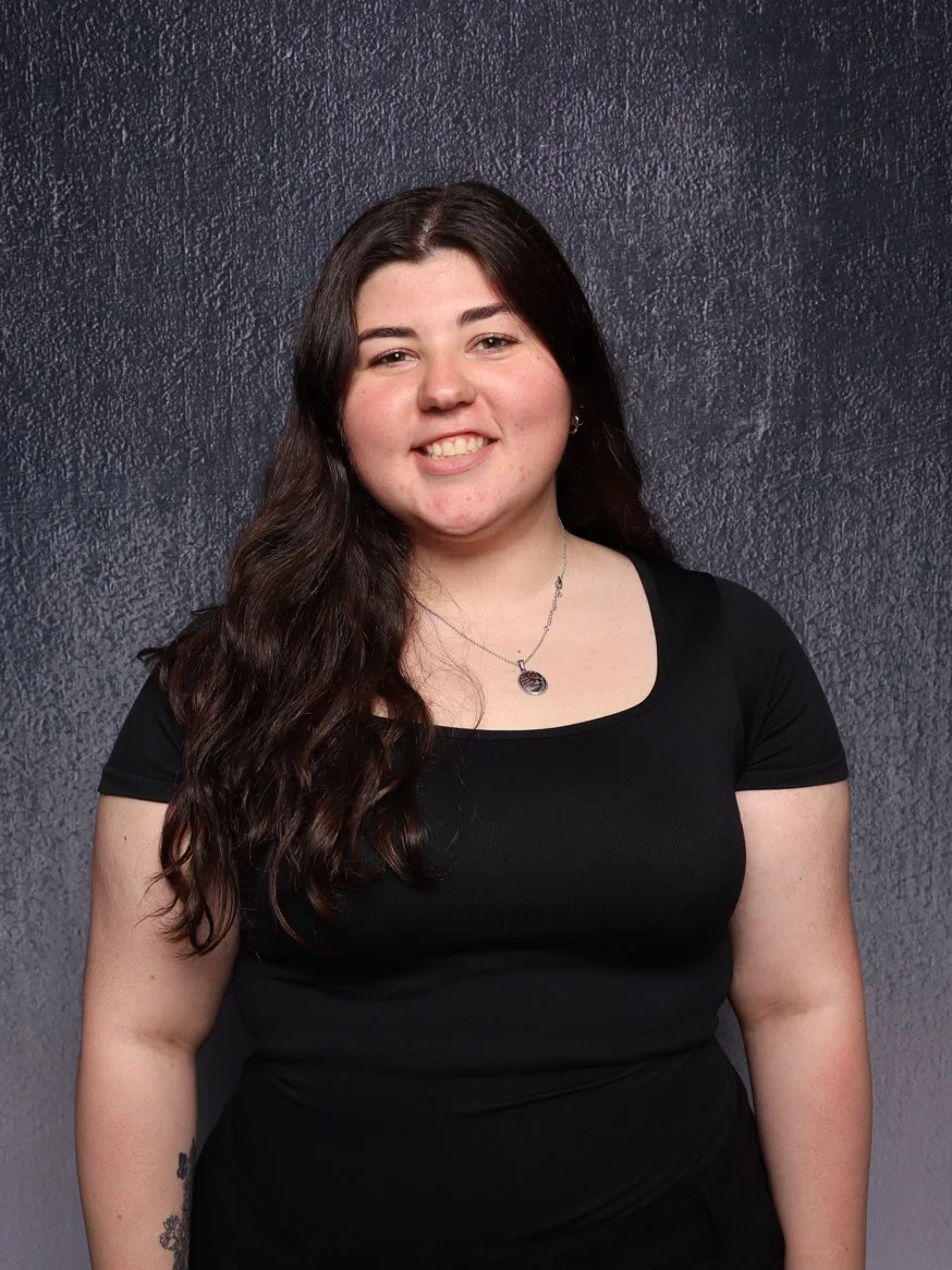 A young woman with long dark hair, wearing a black t-shirt and a silver necklace, smiling while standing against a dark textured background.