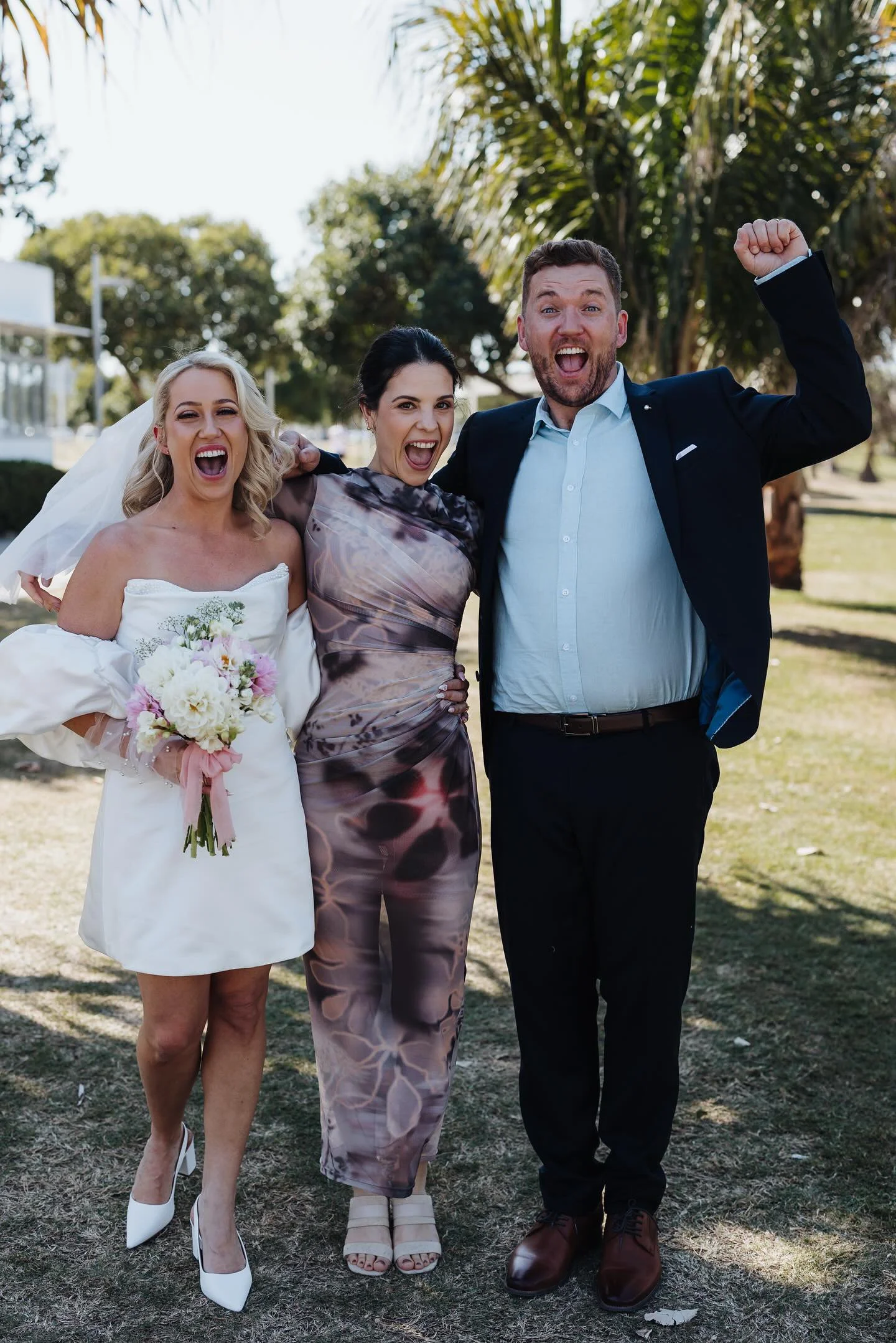 Three people, a bride in a white dress holding a bouquet, and two guests, celebrating outdoors on a sunny day with trees in the background.