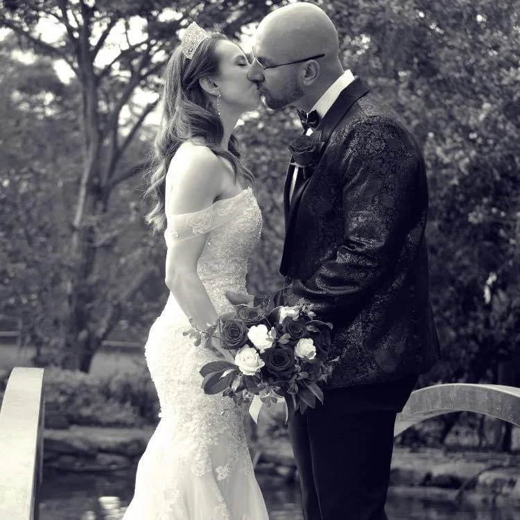 A black and white photo of a bride and groom sharing a kiss outdoors. The bride is wearing an off-shoulder wedding gown and a tiara, holding a bouquet of roses. The groom is wearing a black tuxedo with a bow tie and glasses.