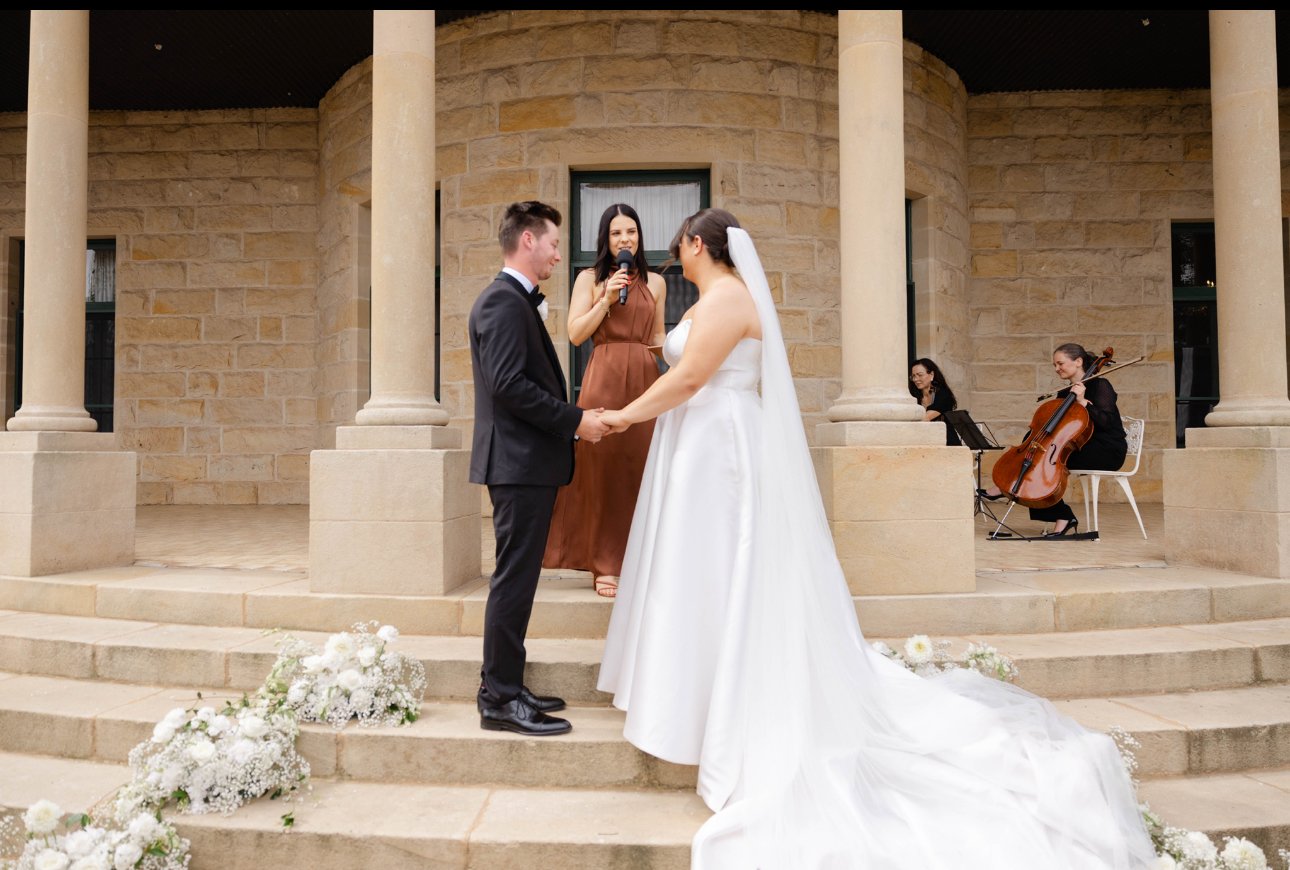A wedding ceremony on the steps of a building with two women and a man. The man and a woman are holding hands and looking at each other. The woman in a white wedding dress has a long train, and the man is in a black tuxedo. A woman in a brown dress is speaking into a microphone. Two women are playing string instruments in the background. Flowers are arranged at the top and bottom of the stairs.