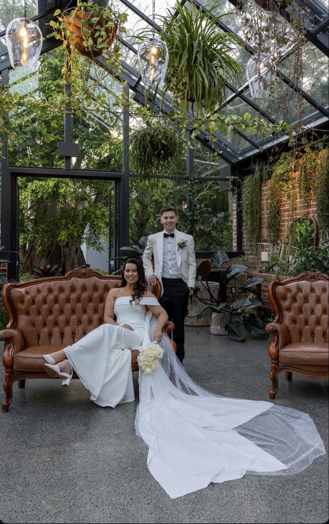 A bride in a white wedding gown sitting on a vintage sofa with a trailing veil, holding a bouquet of white roses, and a groom in a white tuxedo standing beside her, smiling in a lush indoor garden setting with hanging plants and glass ceiling panels.