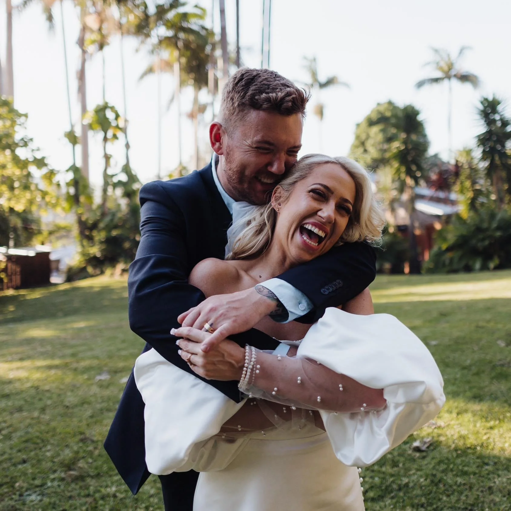A happy couple, the man in a black suit and the woman in a white wedding dress, joyfully hugging outdoors with palm trees in the background.