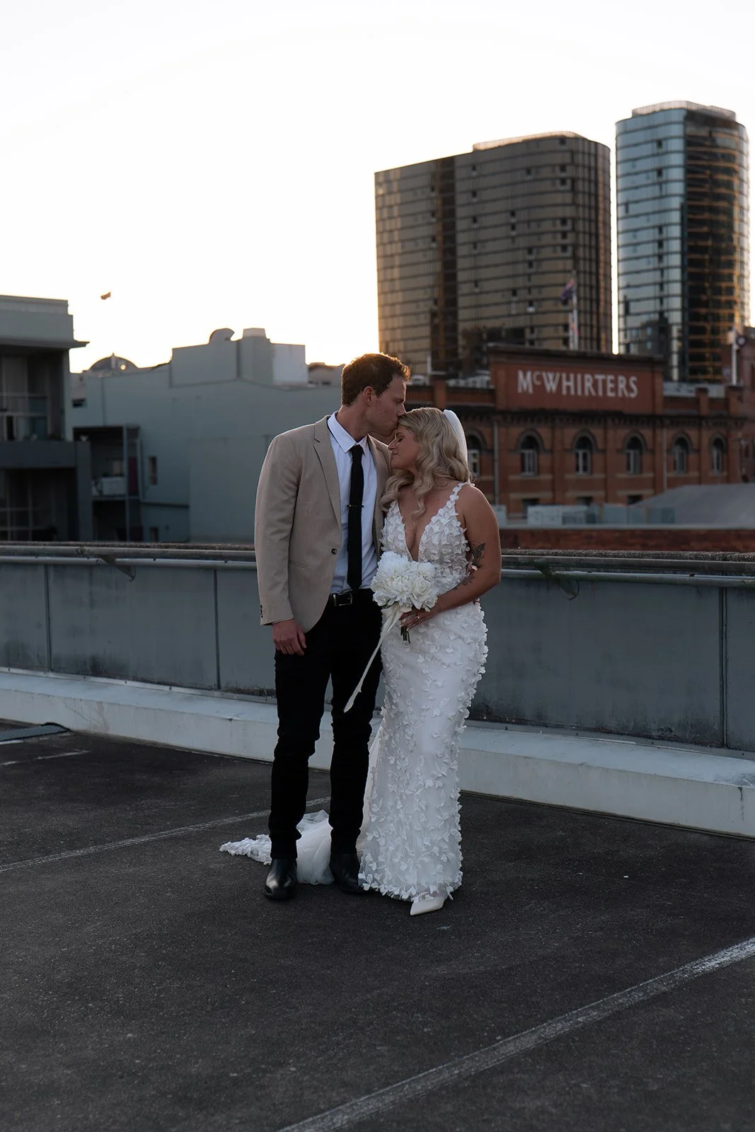 A bride and groom stand close together on a parking garage rooftop during sunset, with city buildings in the background. The groom leans his forehead against the bride's forehead, and she holds a bouquet of white flowers.