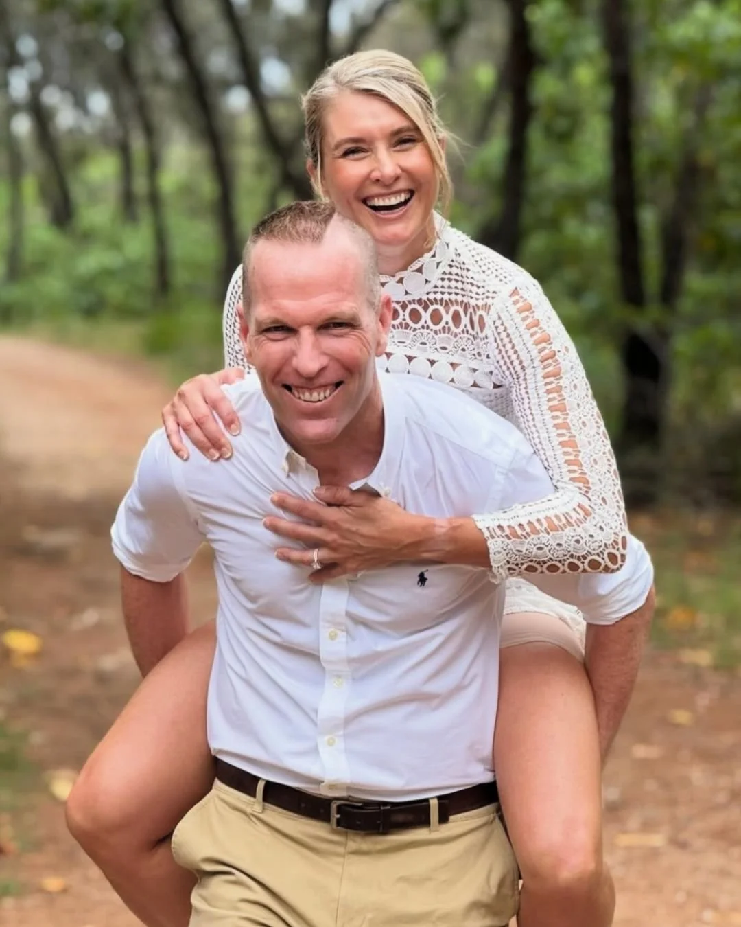 A smiling couple outdoors in a forest, with the woman on the man's back, both wearing light-colored clothing.