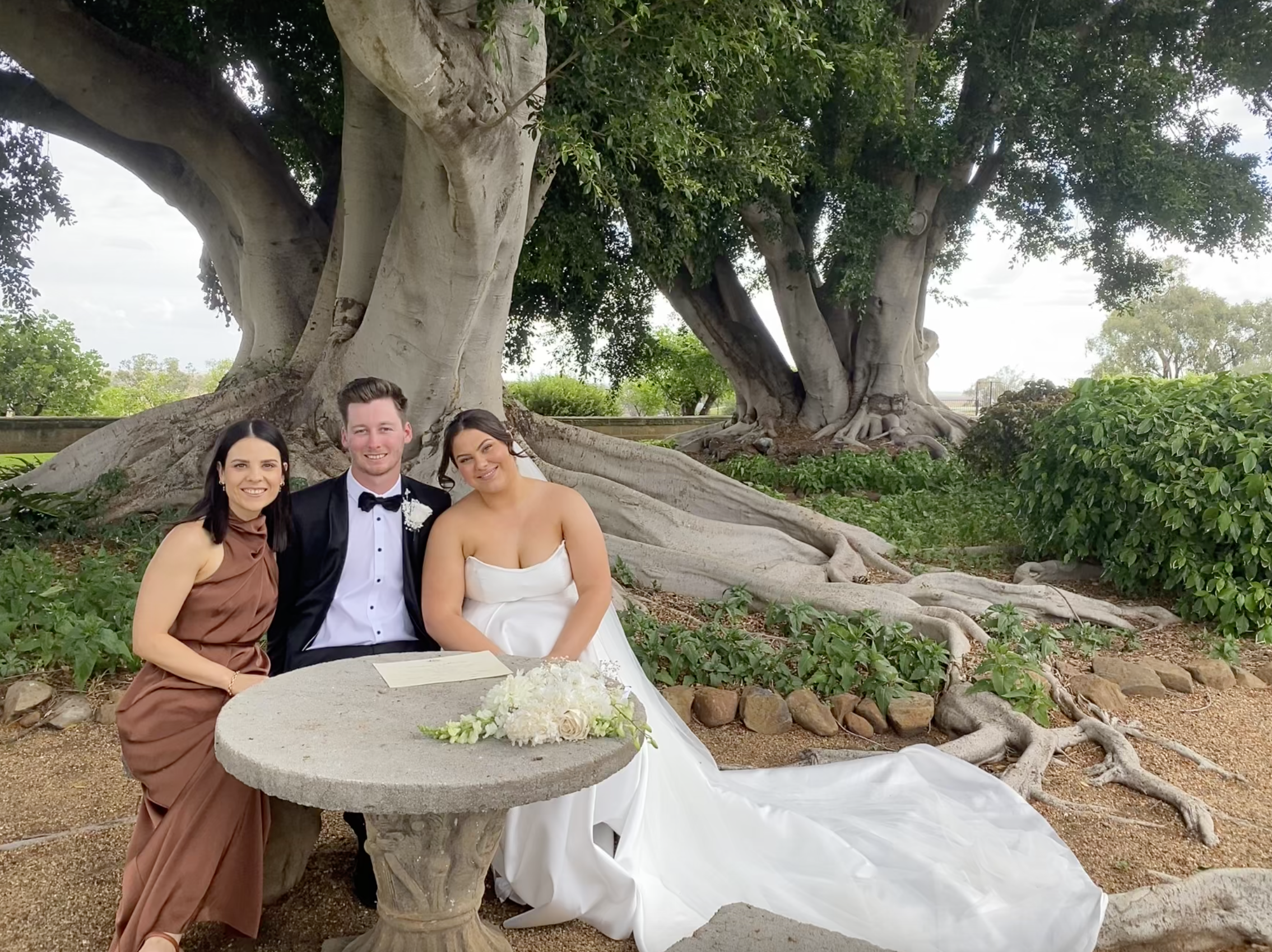 A wedding scene with three people sitting at a stone table outdoors, with a large tree in the background. The person in the middle is wearing a tuxedo, and the person on the right is wearing a white wedding dress. The person on the left is wearing a brown dress. There is a bouquet of white flowers on the table.