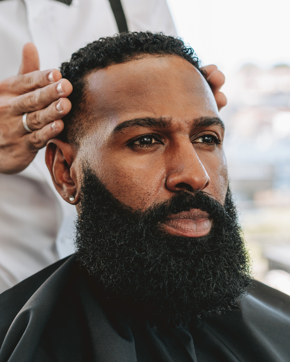 A man with a thick beard getting a haircut in a barbershop, wearing a black cape, with a focused expression as a barber touches his hair.