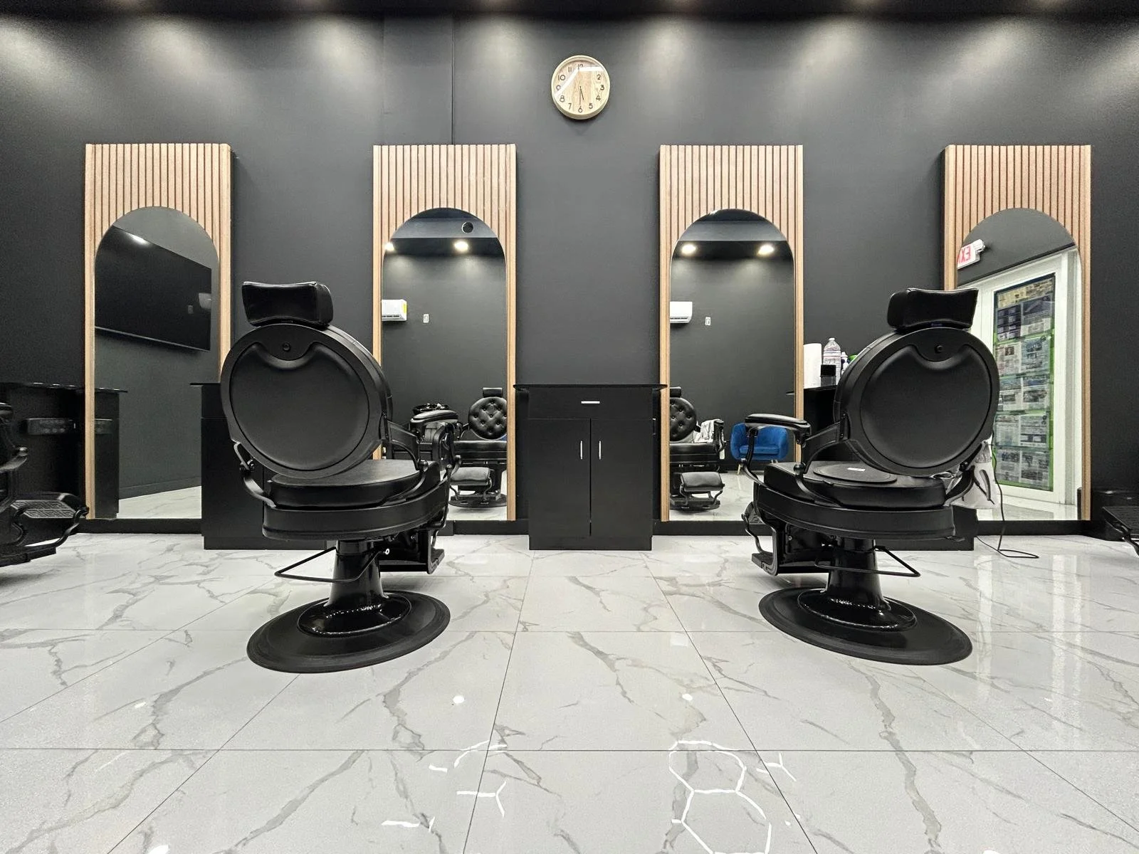 A modern barbershop, Platinum Cuts, with two black barber chairs facing large mirrors on a black wall. There is a clock above the middle mirror, and a small black cabinet between the chairs. The floor is made of white marble with grey veining.