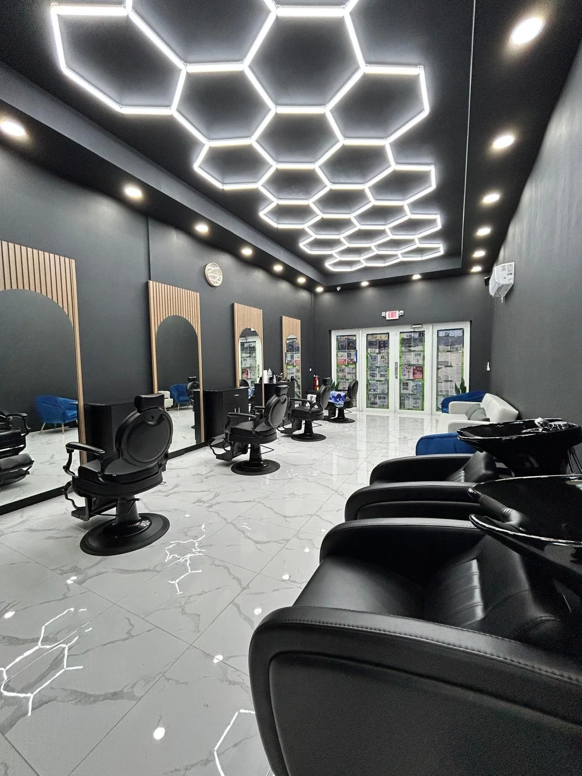 Modern barbershop, Platinum Cuts, with black chairs, mirrors, sink basins, and sleek white marble tile flooring, illuminated by geometric hexagon ceiling lights.