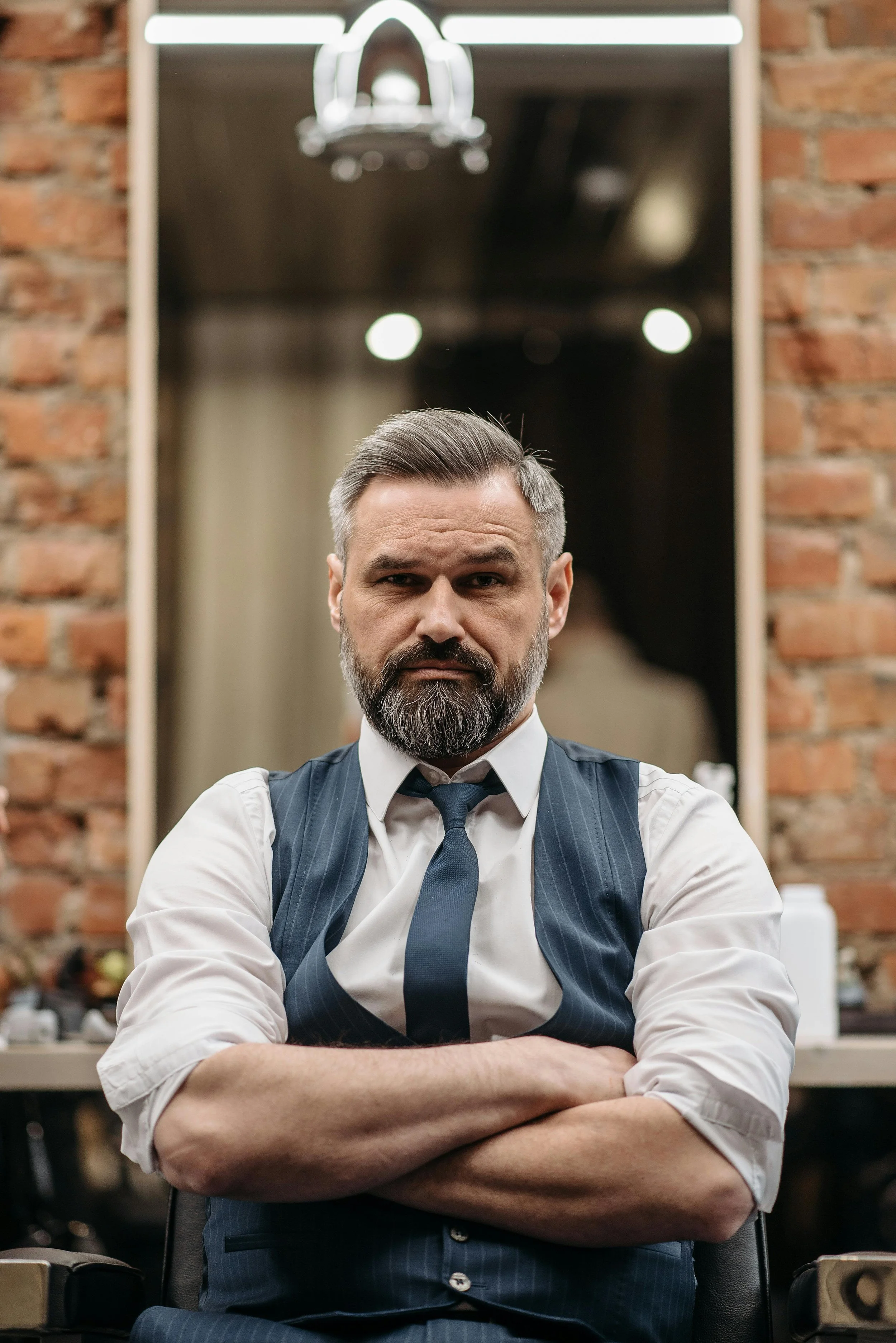 A confident man with a beard and slicked-back hair standing with arms crossed in front of a mirror, in a stylish room with exposed brick walls.