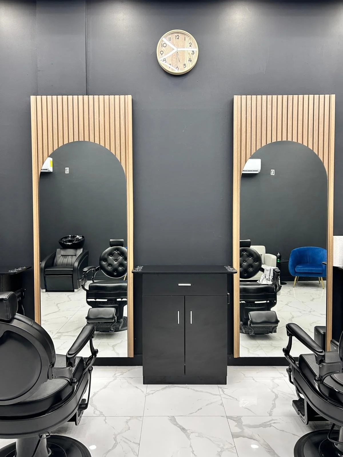 Hair salon with two styling stations, each with large mirrors, black chairs, and wooden paneling. A clock shows approximately 10:10. The floor is marble, and there is a black cabinet between the stations. Additional salon chairs and a blue armchair a