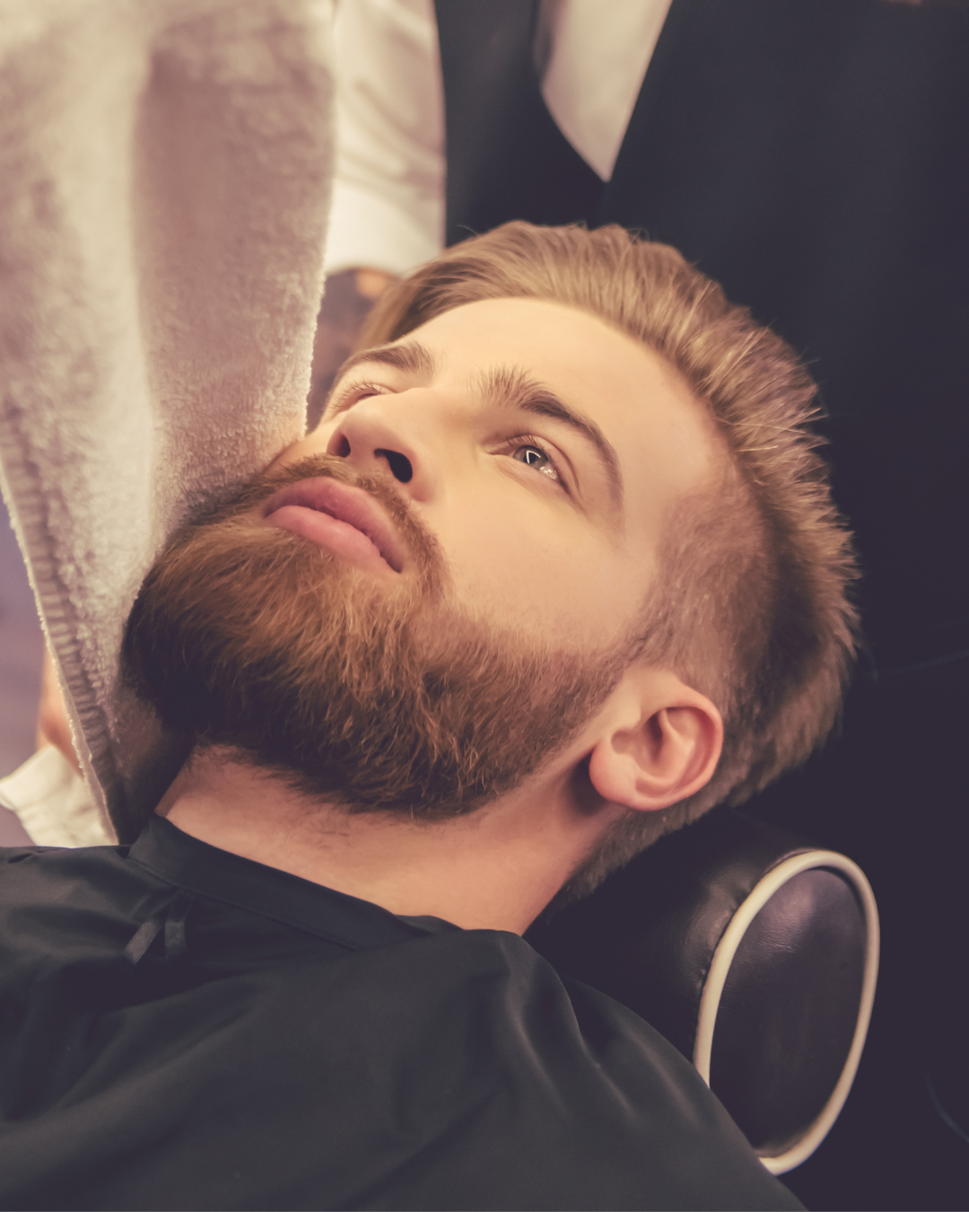 A man with a beard and mustache lying on a chair in a barbershop, gazing upwards.