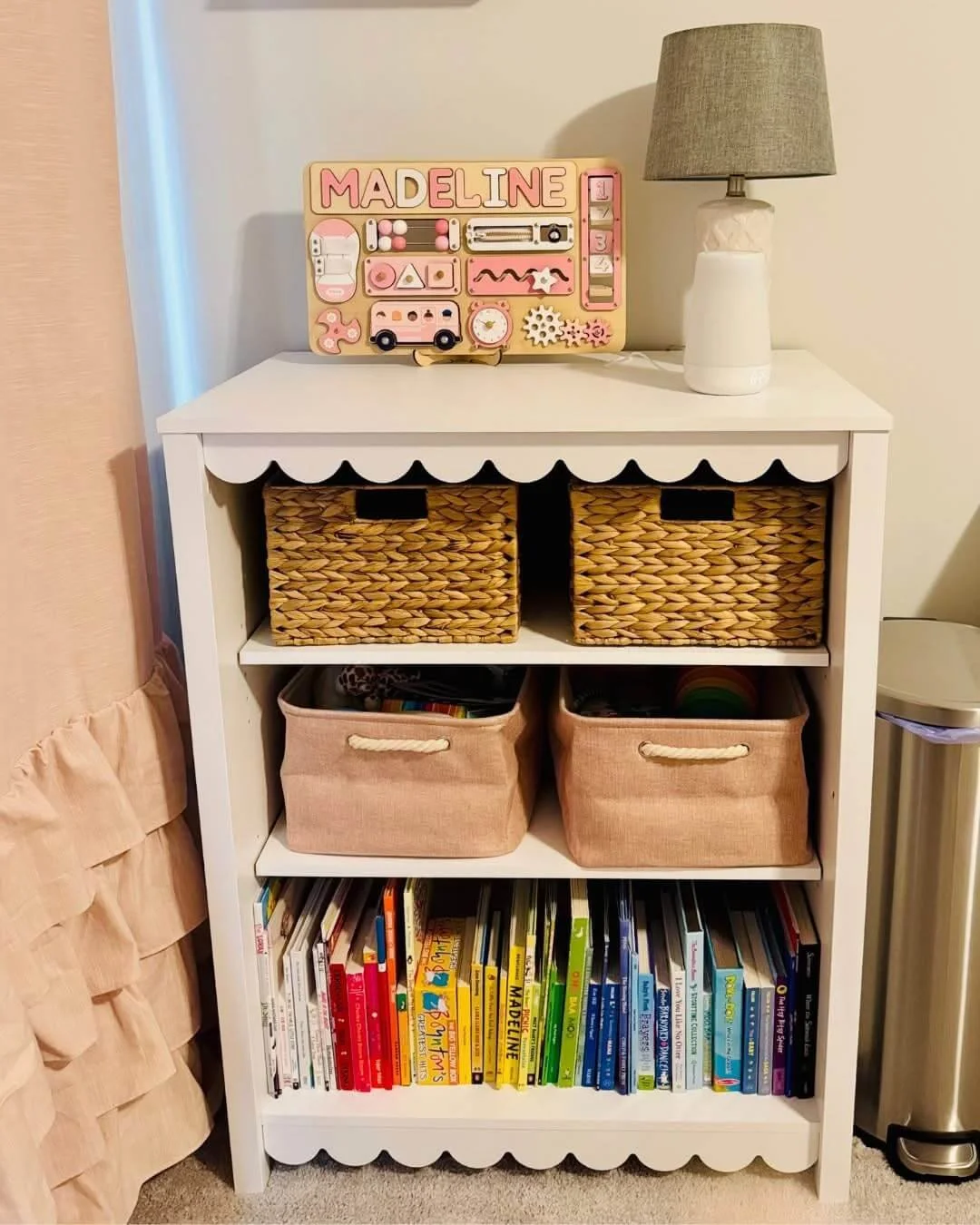 White bookshelf with a top section holding a pink and cream nameplate displaying 'MADELINE' in pink, along with various educational toys, and a lamp with a beige shade. The middle section contains colorful children's books arranged vertically, and the bottom section holds woven baskets and other storage bins. A trash can with a lid is visible to the right of the bookshelf.