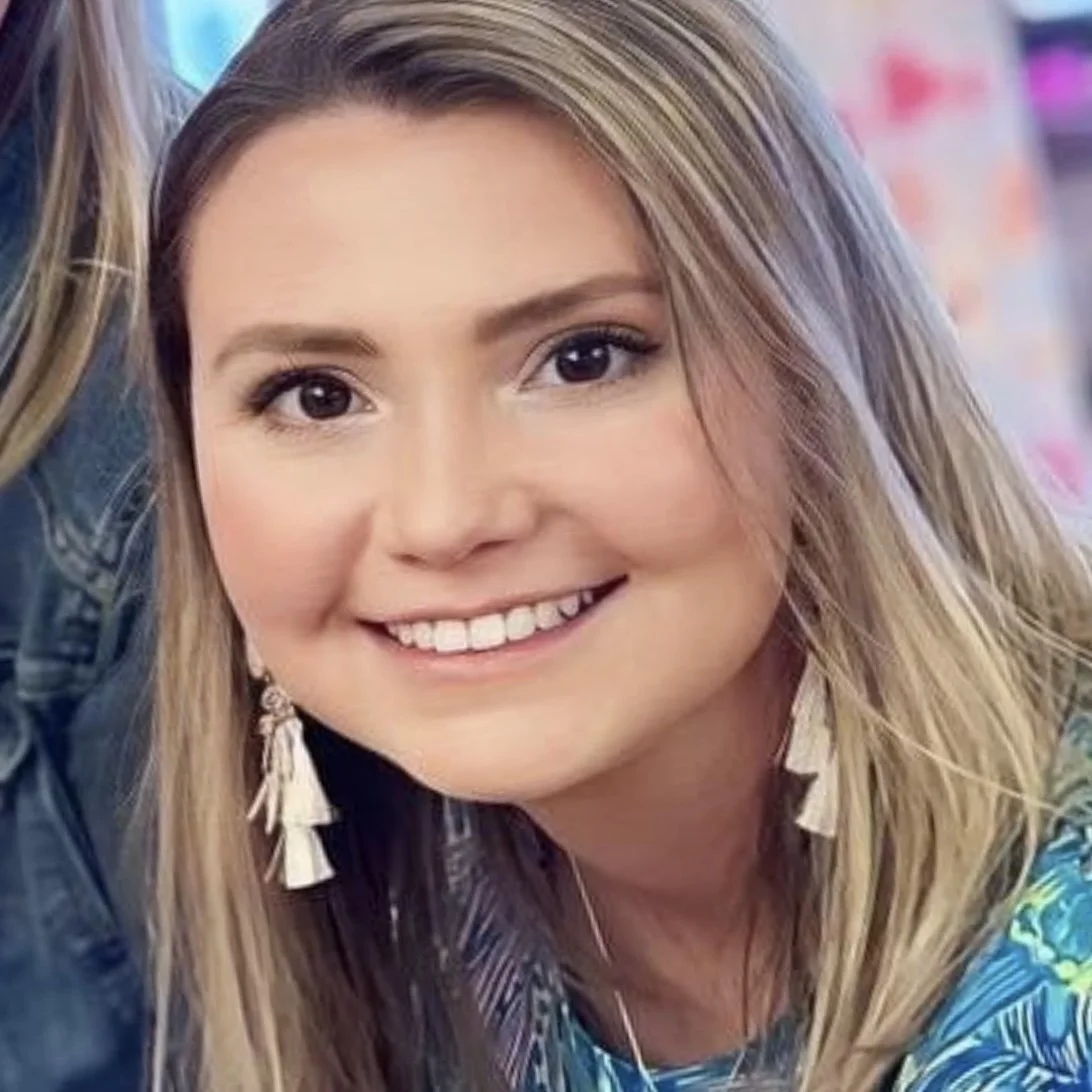 Close-up of a young woman smiling with blonde hair, wearing earrings and a colorful top.
