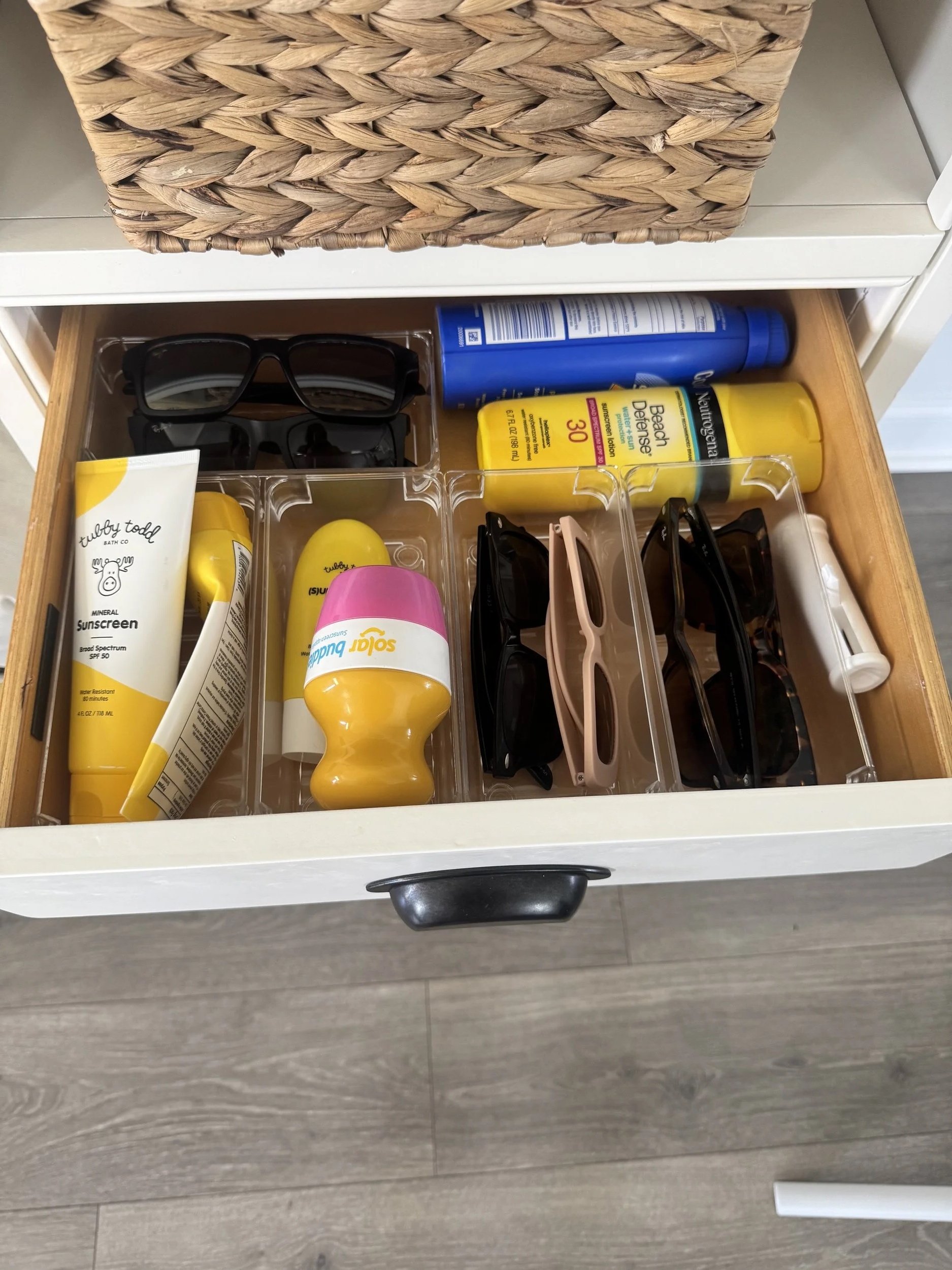 Open drawer containing black sunglasses, a yellow sunscreen bottle, a pink sunscreen bottle, a blue spray bottle, and several smaller items.