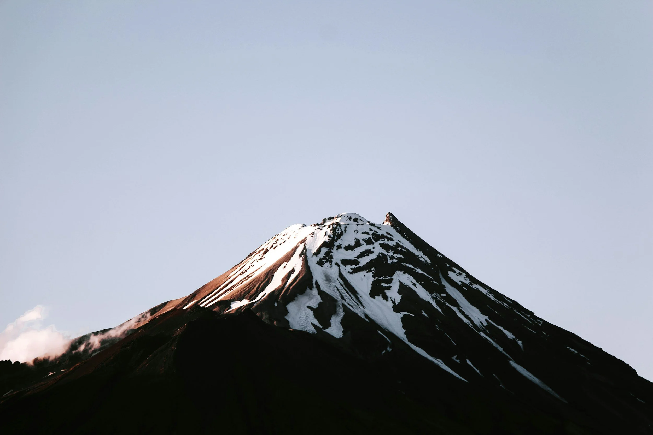 Snow-capped volcanic mountain with dark slopes and a light sky background.