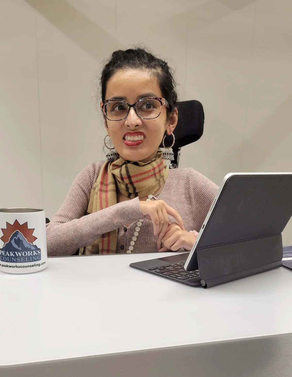 A woman with glasses making a grimace while sitting at a desk with a tablet and a mug that has a mountain logo and the words "PEAKWORKS COUNSELING" on it.