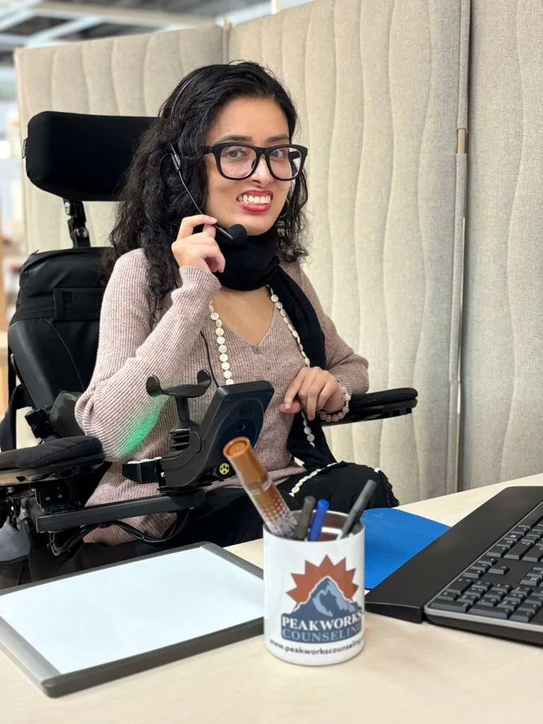 A woman with dark curly hair, glasses, and a headset, sitting in a wheelchair at a desk with a laptop and a container holding pens and a thermometer, in an office setting.
