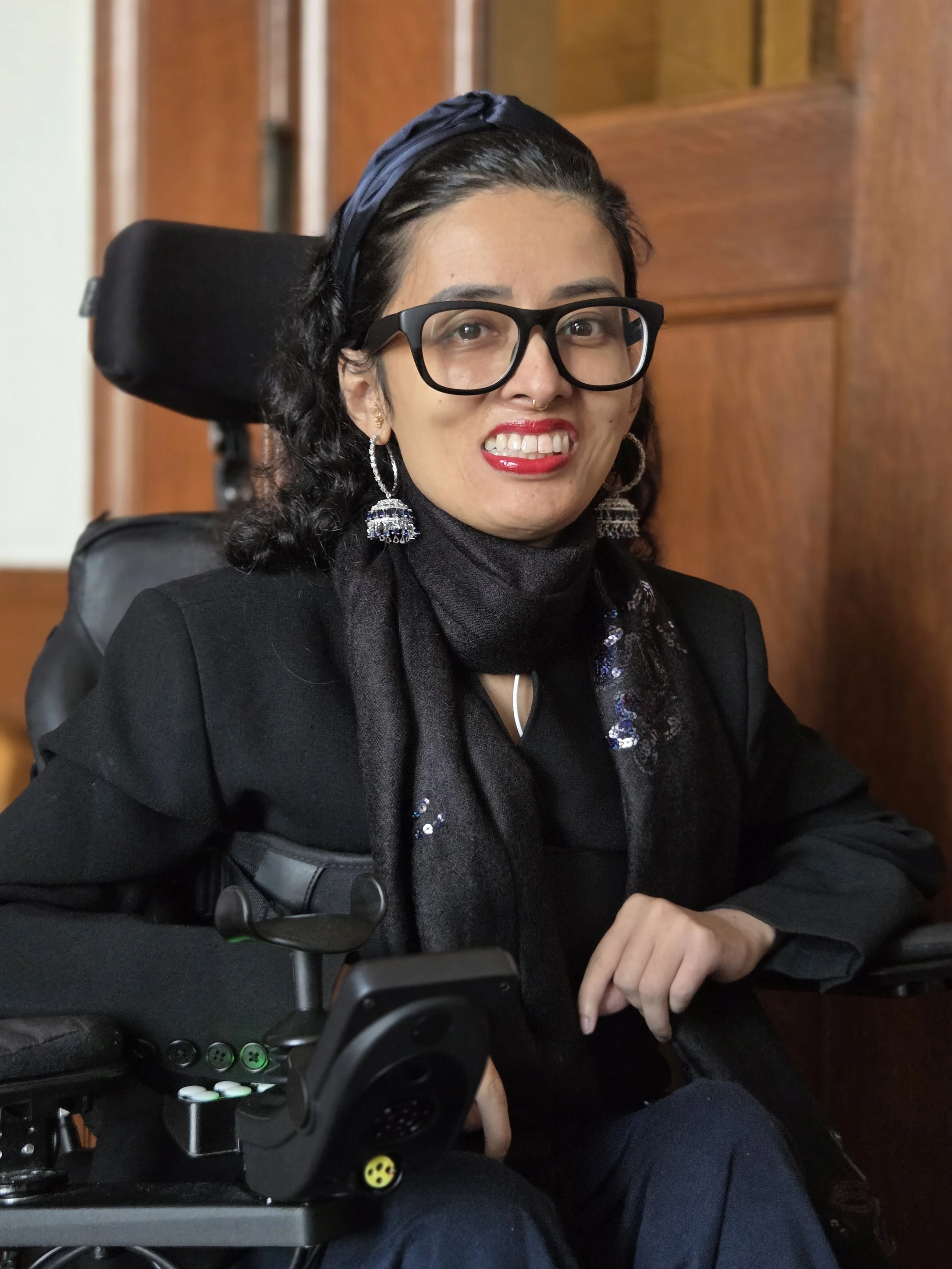 A woman with dark, curly hair, wearing large black glasses, earrings, and a headband, sitting in a wheelchair with a wooden background behind her.
