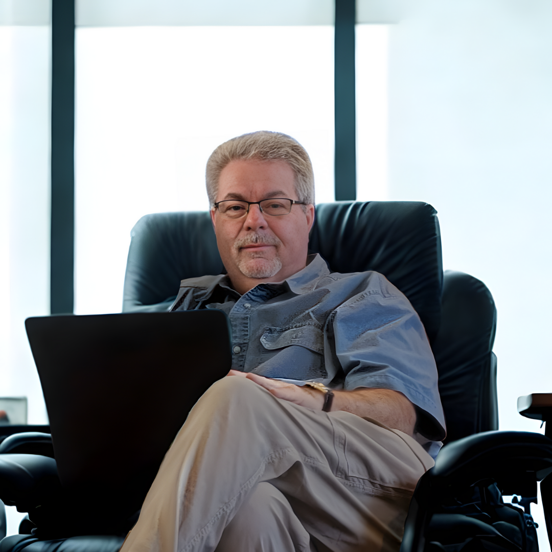 Lyle Chinkin, air quality expert, a man with glasses and gray hair sits in a black leather office chair with a laptop on his lap, in front of large windows in a modern office.