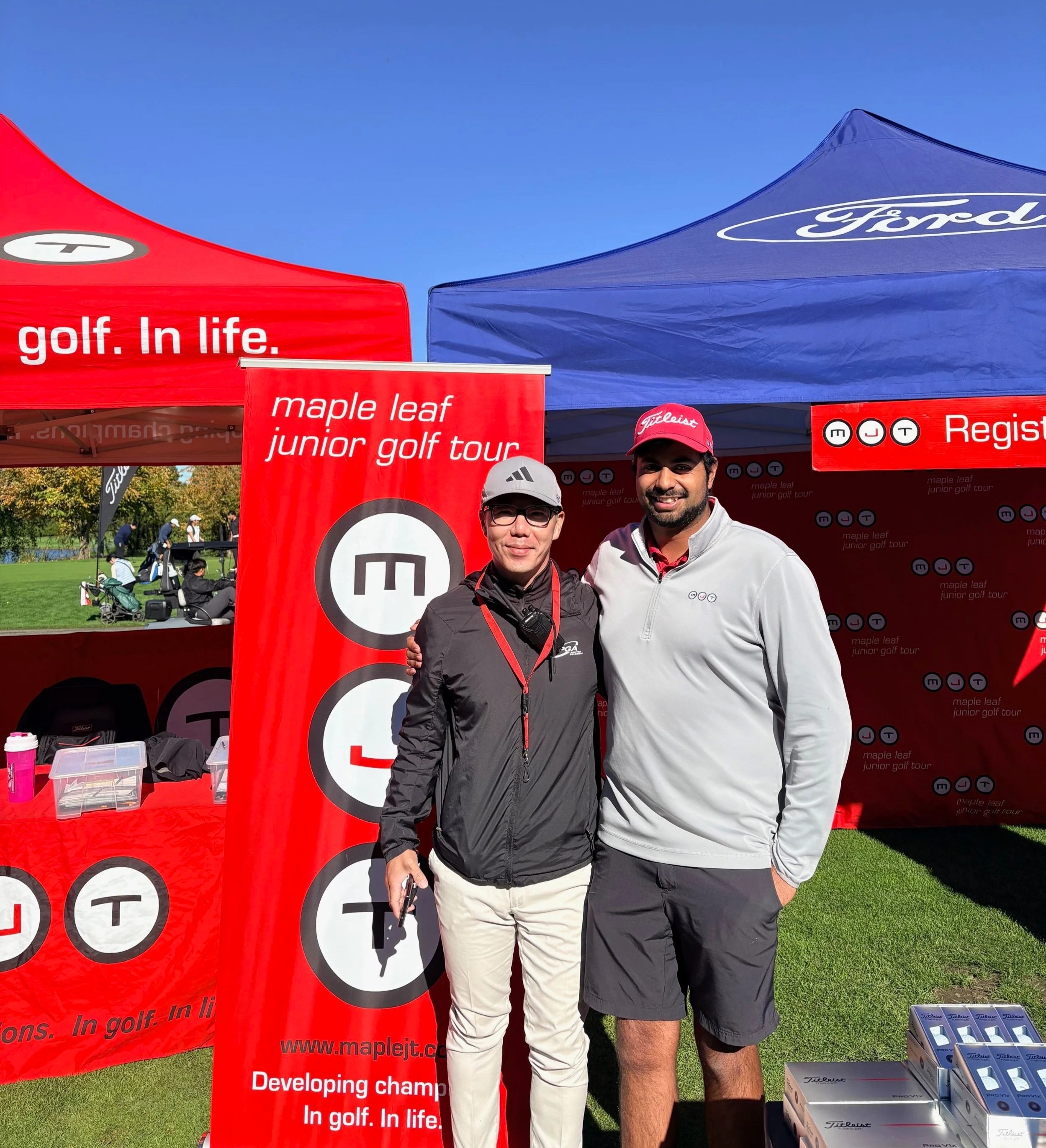 Two men standing together outdoors at a golf event, smiling for the photo. They are in front of tents with branding from the Maple Leaf Junior Golf Tour and other golf-related items, including boxes of golf clubs or equipment.