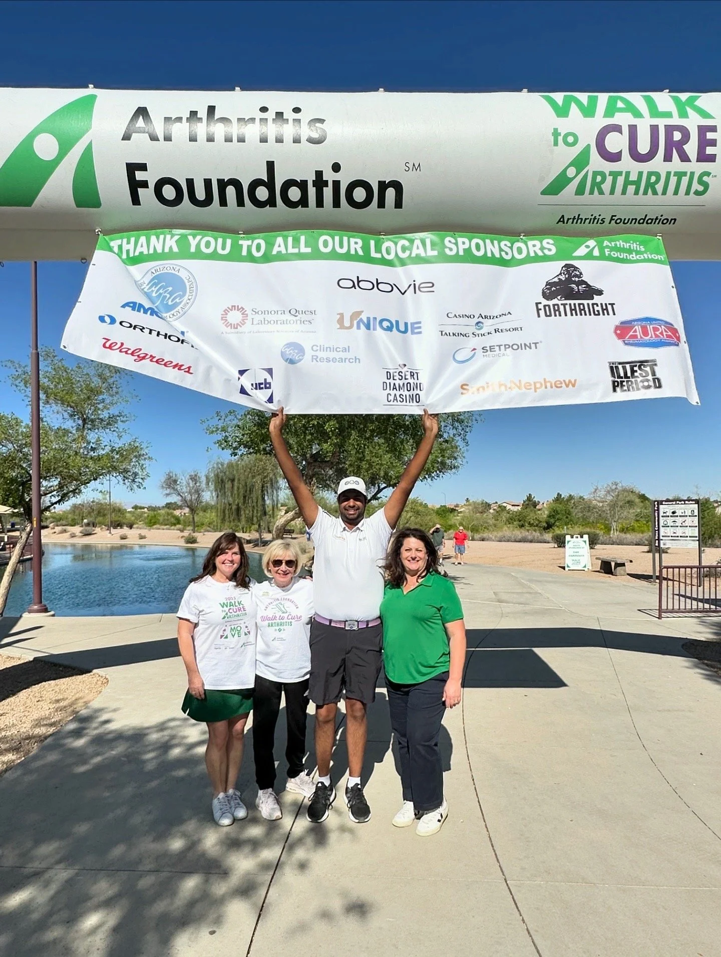 Group of four smiling people standing outdoors in front of a lake and trees, holding a large banner that promotes arthritis awareness and charity walk event.