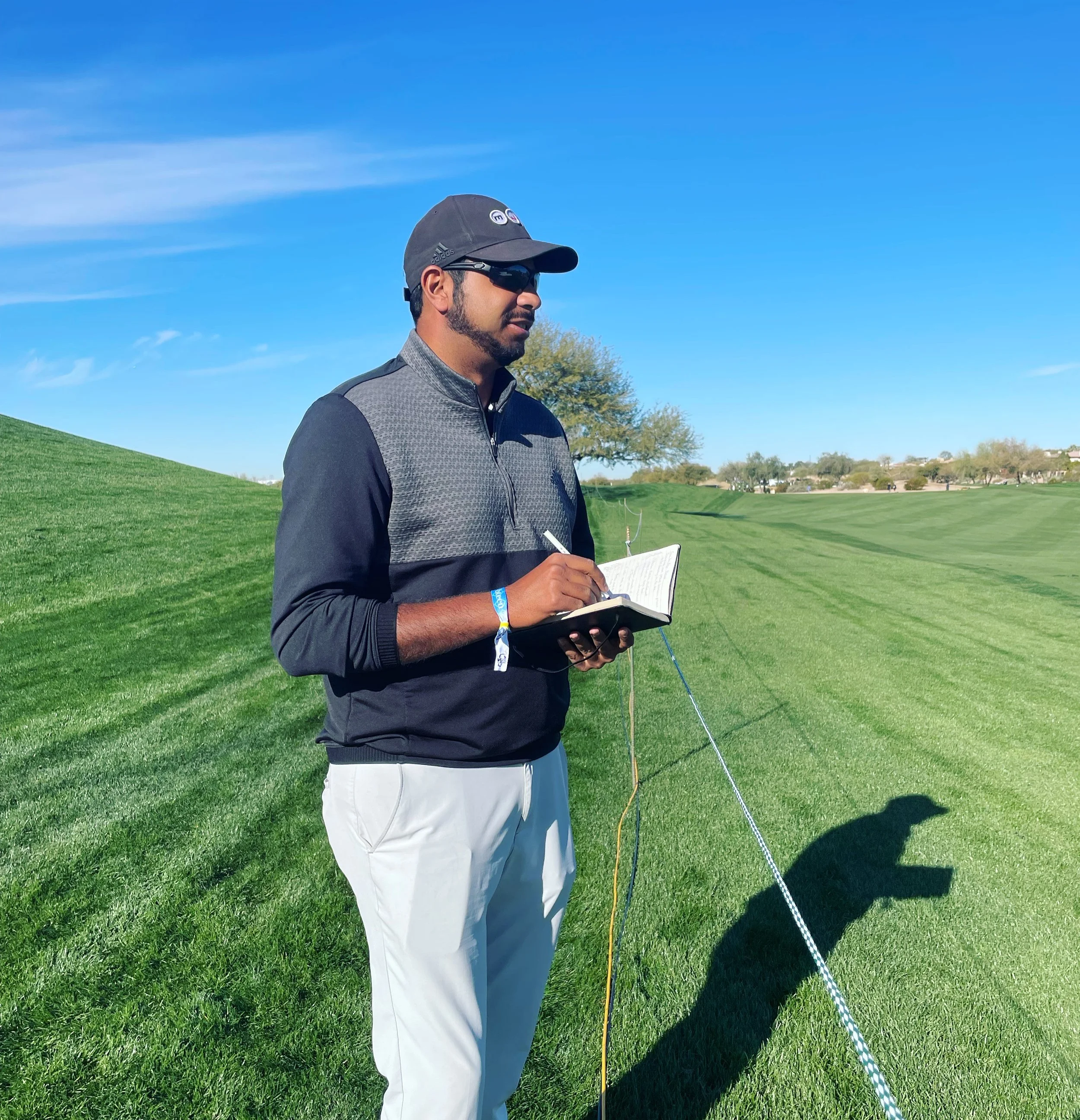 Man standing on a golf course holding a notepad and pen, wearing sunglasses, a hat, and a golf jacket, with a green field, trees, and blue sky in the background.