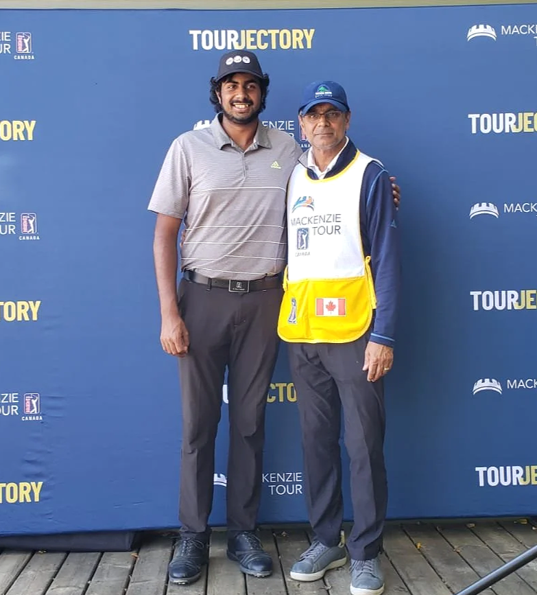 Two men standing in front of a blue backdrop with logos, one wearing a gray polo shirt and the other wearing a jacket and a bib with a Canadian flag, at the Mackenzie Tour.