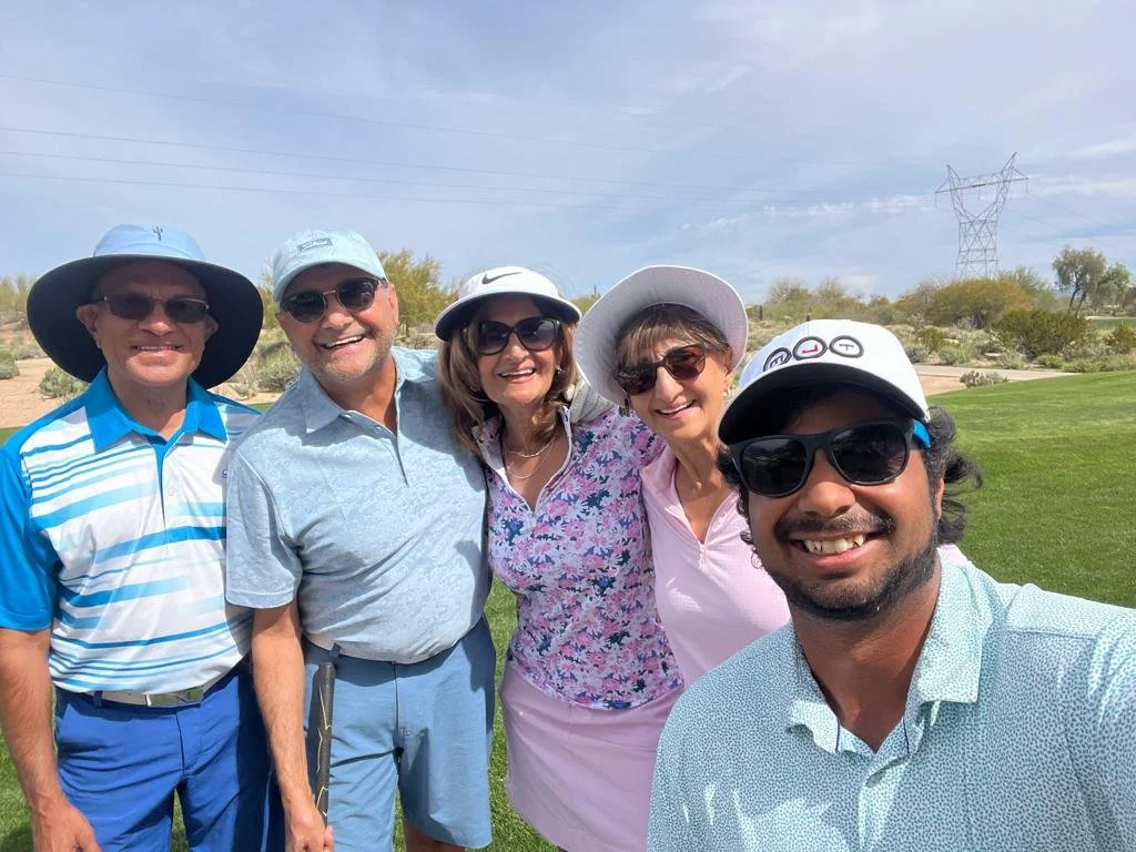 Group of five people smiling and posing for a selfie on a golf course, wearing hats and sunglasses.