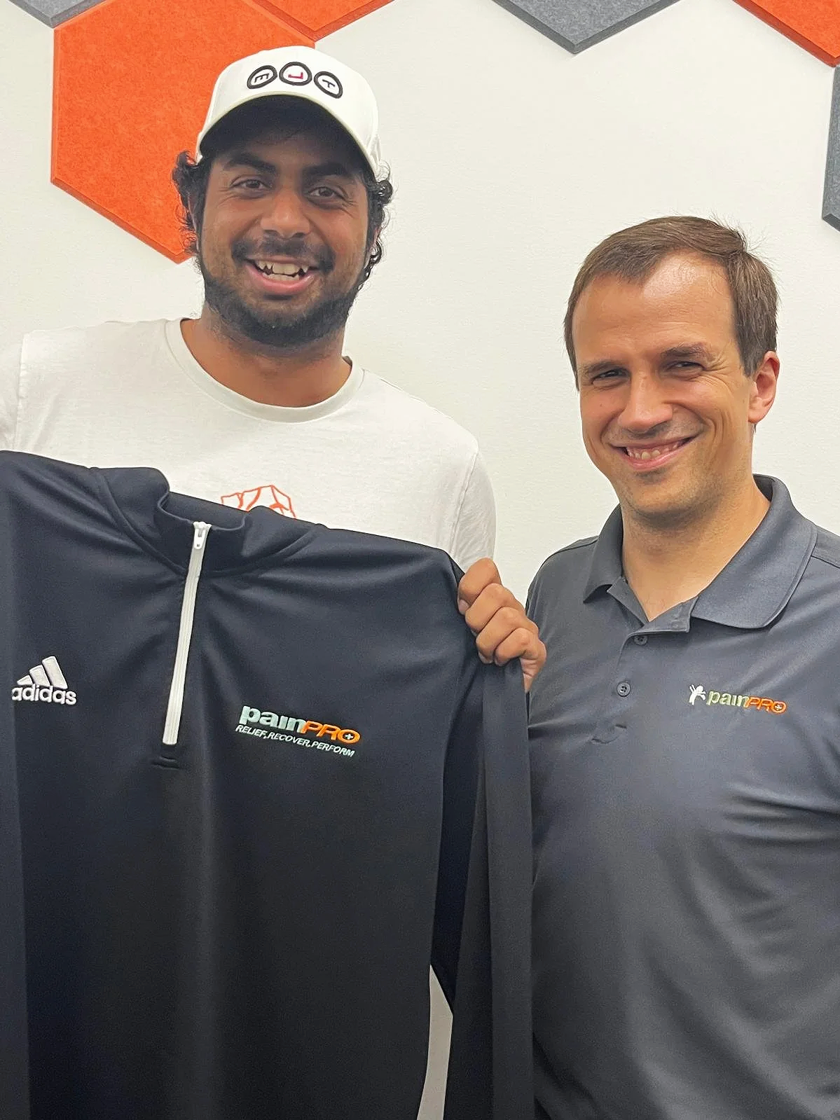 Two men smiling and holding a black shirt with the 'painPRO' logo, standing indoors against a white wall with orange and black acoustic panels.
