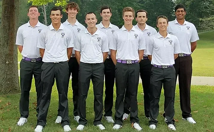 Eight young men in white polo shirts and dark pants stand outdoors on grass, smiling for a group photo with trees and a sports field in the background.