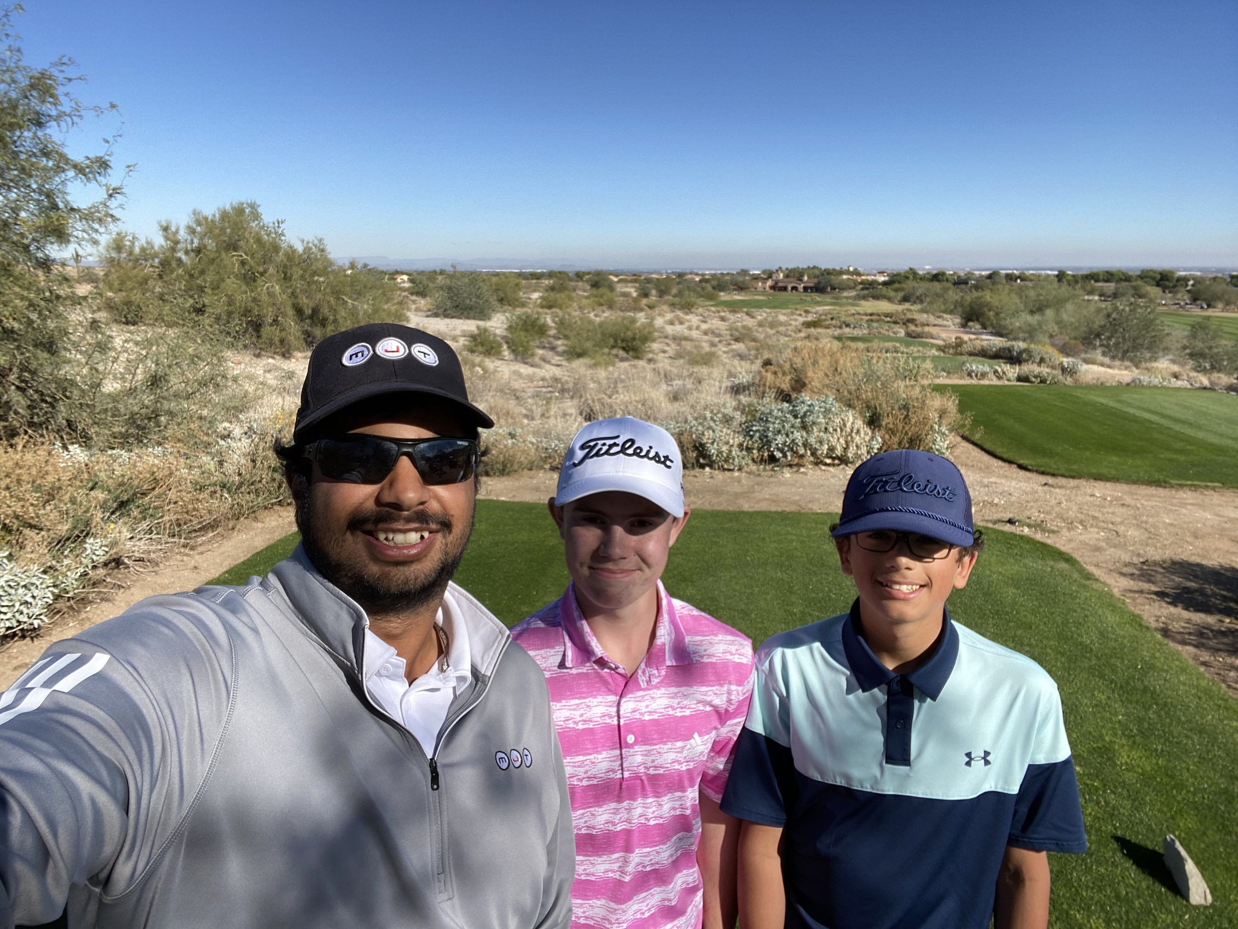 Three males standing on a golf course with desert landscape and blue sky in the background.