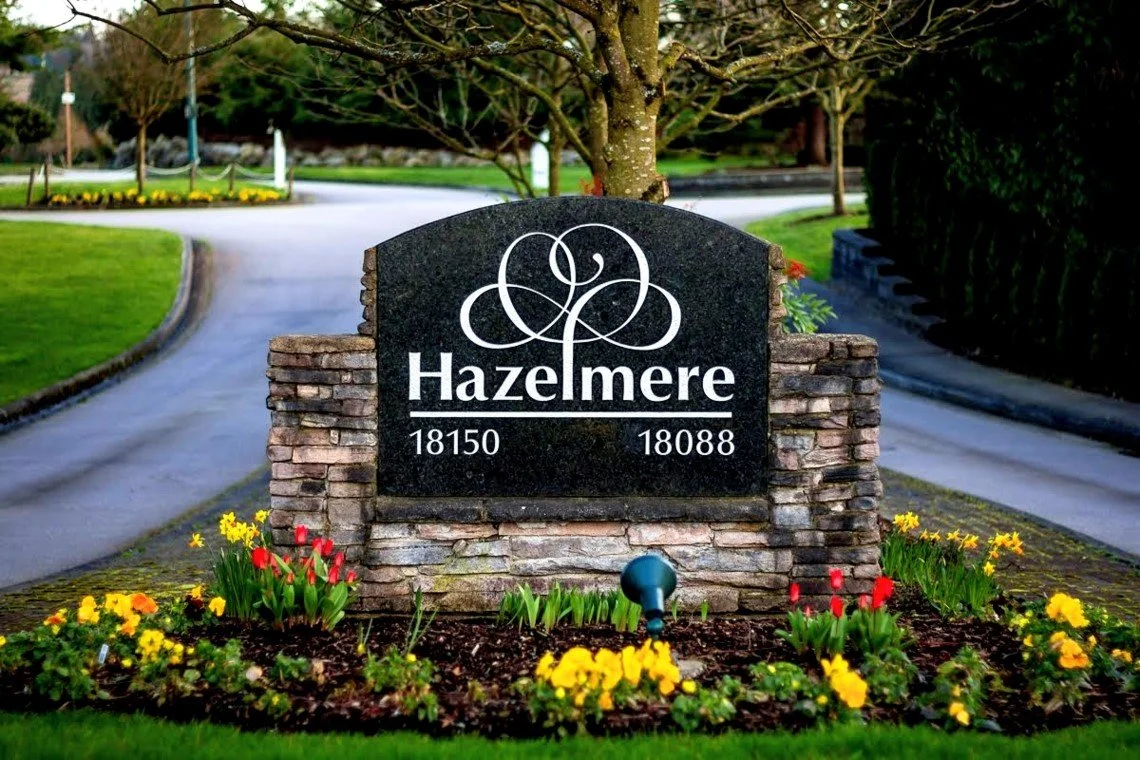 A stone and brick monument with the name Hazelmere and addresses 18150 and 18088, surrounded by colorful flowers in a well-maintained garden, with a winding road and trees in the background.