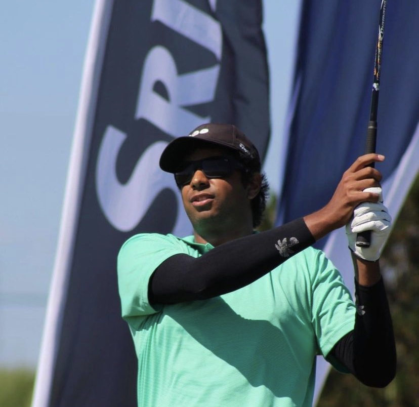 A young man wearing a black cap, sunglasses, and a mint green shirt holding a golf club, standing outdoors.