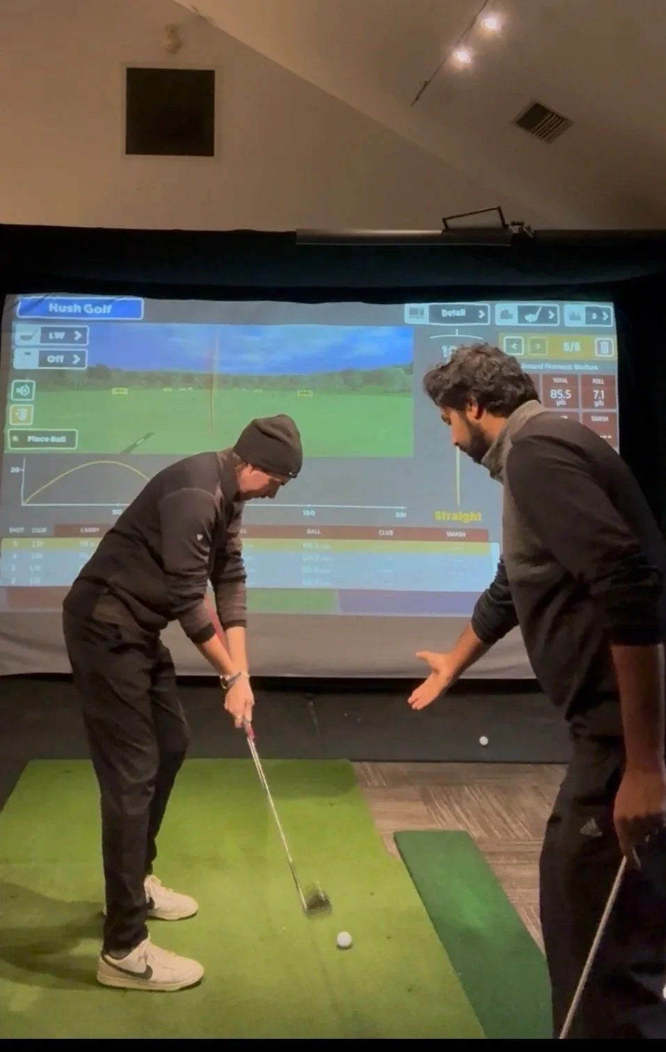 Two men playing indoor golf on a simulation screen. One man is preparing to hit a golf ball with a club, while the other man is observing and pointing. The golf coach is guiding the player's golf swing.