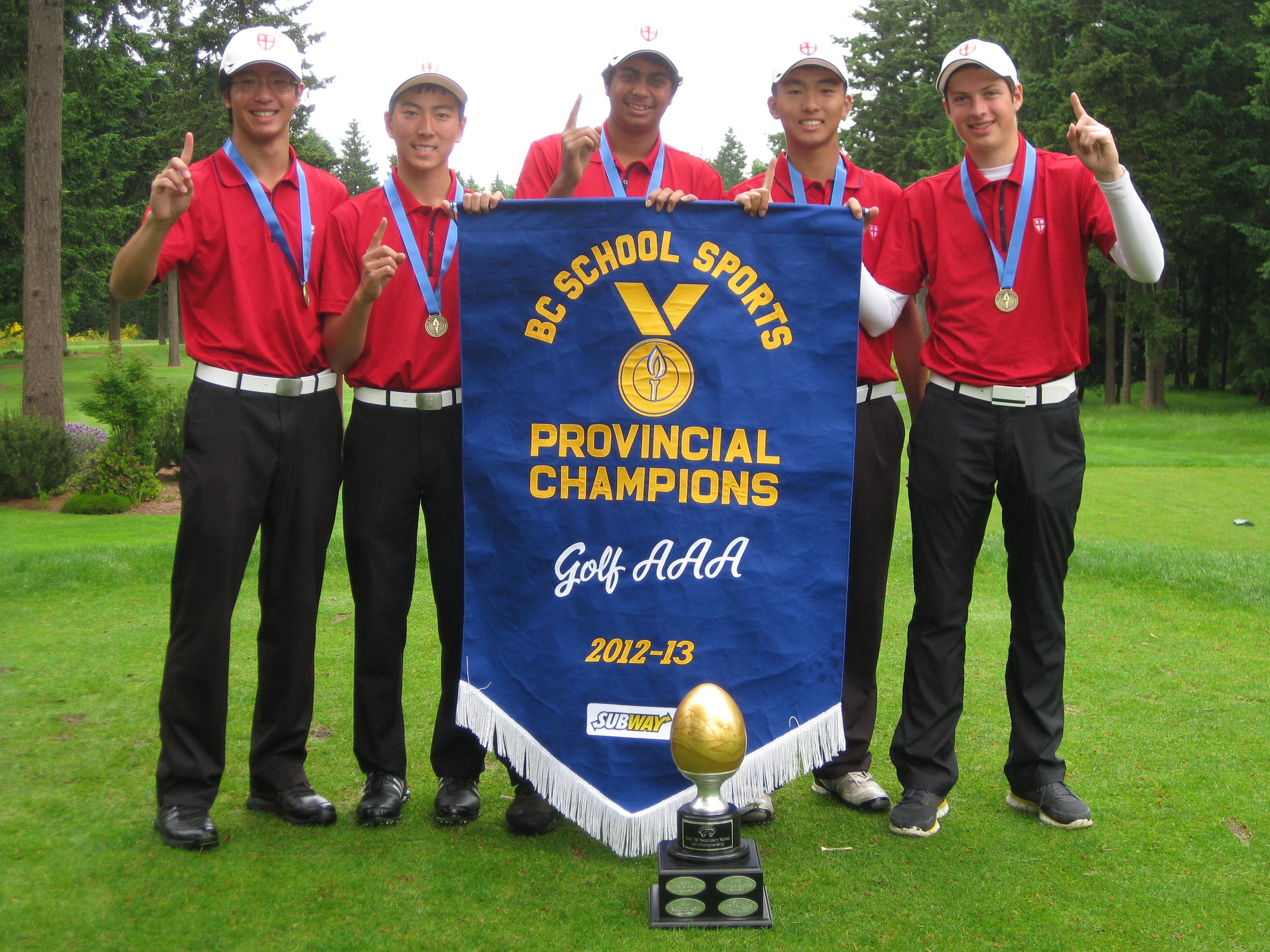 Group of six young male golfers in red shirts and black pants with medals around their necks, displaying a blue banner that reads 'BC School Sports Provincial Champions Golf AAA 2012-13' on a golf course with trees in the background.